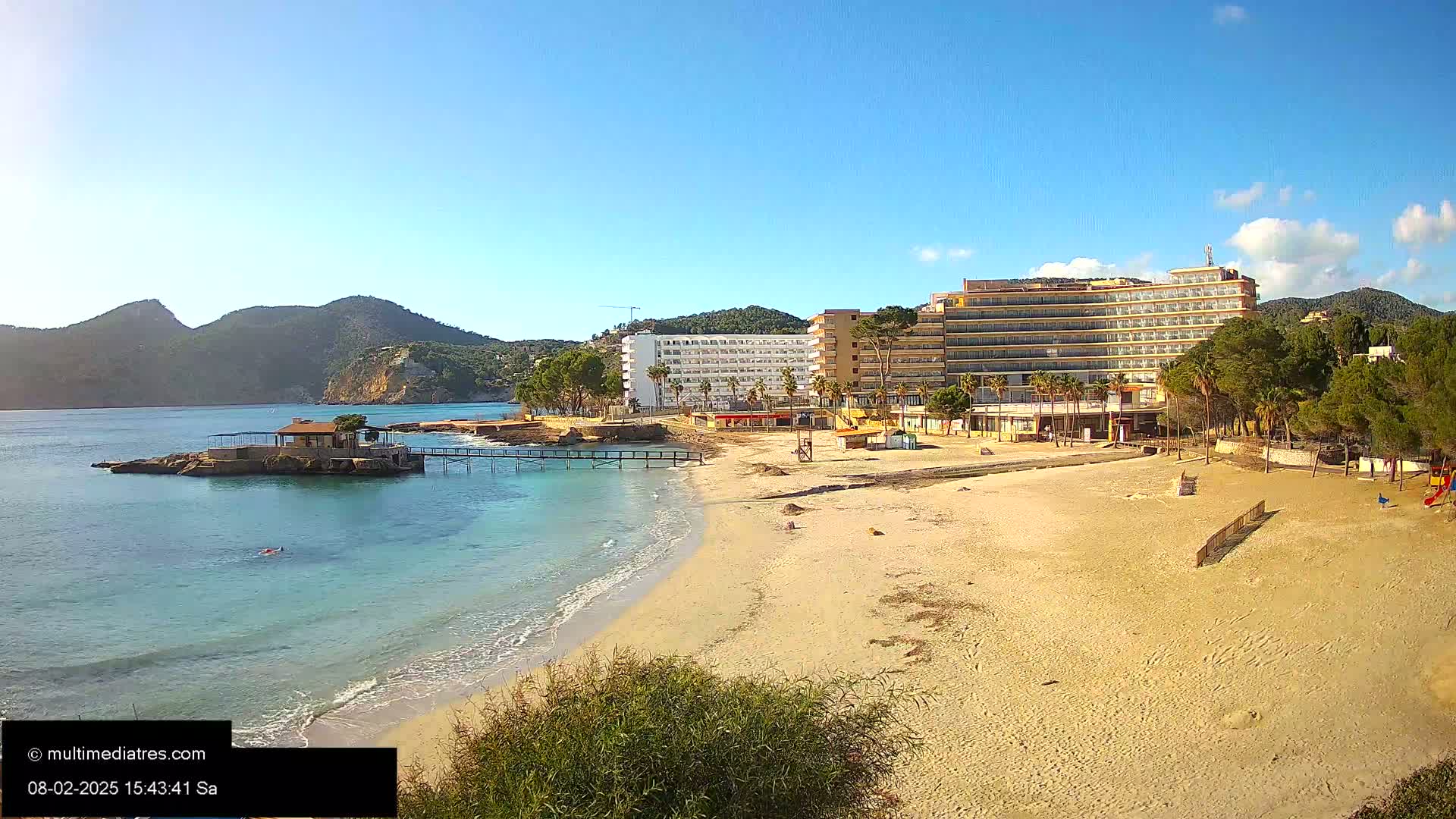 A sandy beach with calm, clear water laps at its shore, backed by a large hotel and a small island with a structure on it, all under a bright, sunny sky.