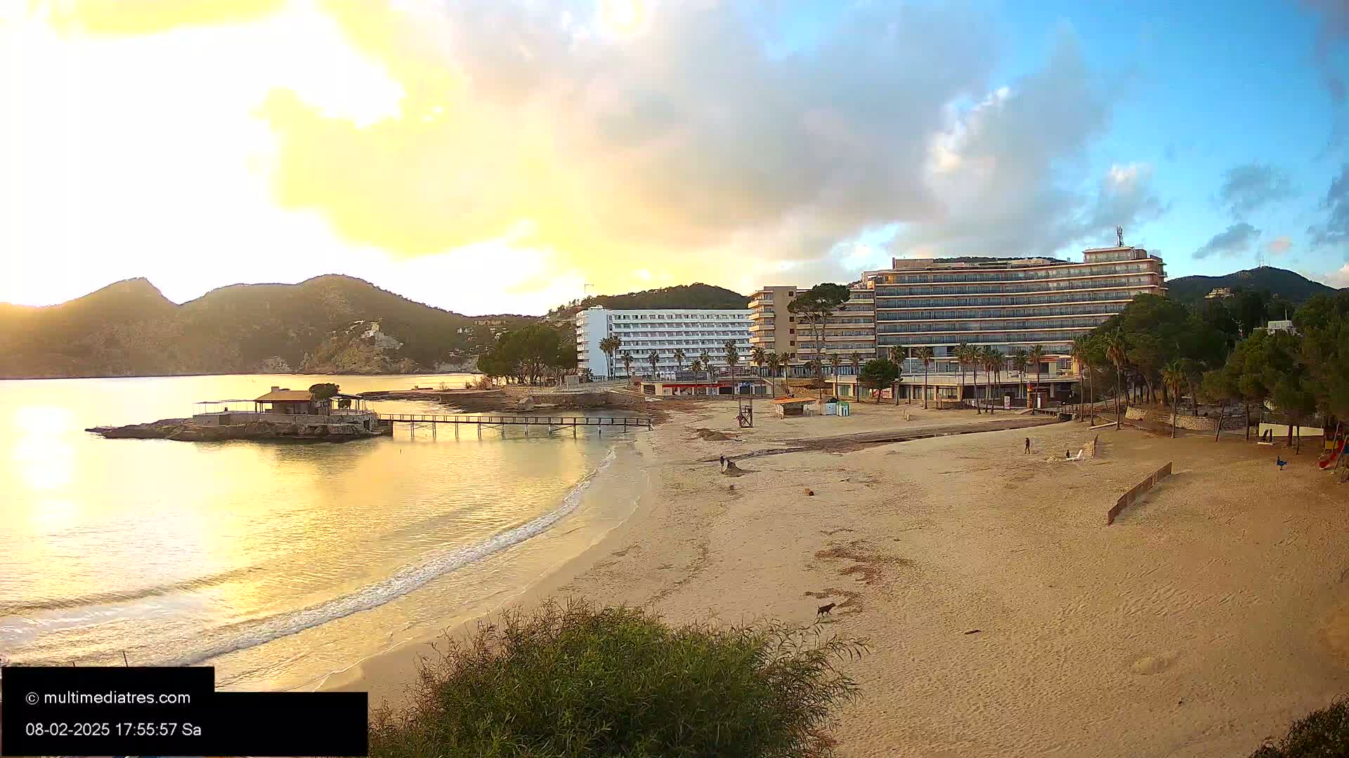 A sandy beach at sunset with calm water, a small pier, a beachfront restaurant, and several hotels in the background.