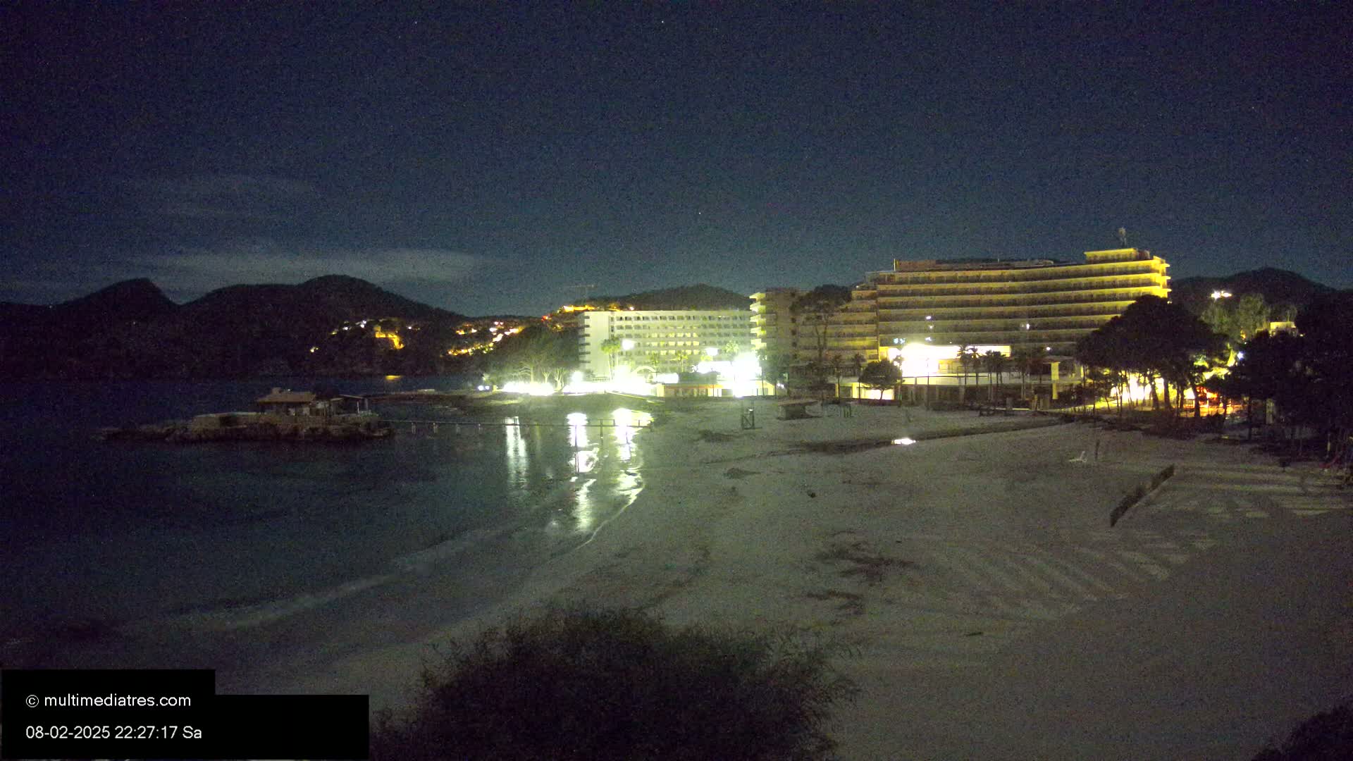 A nighttime view of a beach with two illuminated hotels and a small building on a rocky outcropping in the calm water, under a clear, starry sky.