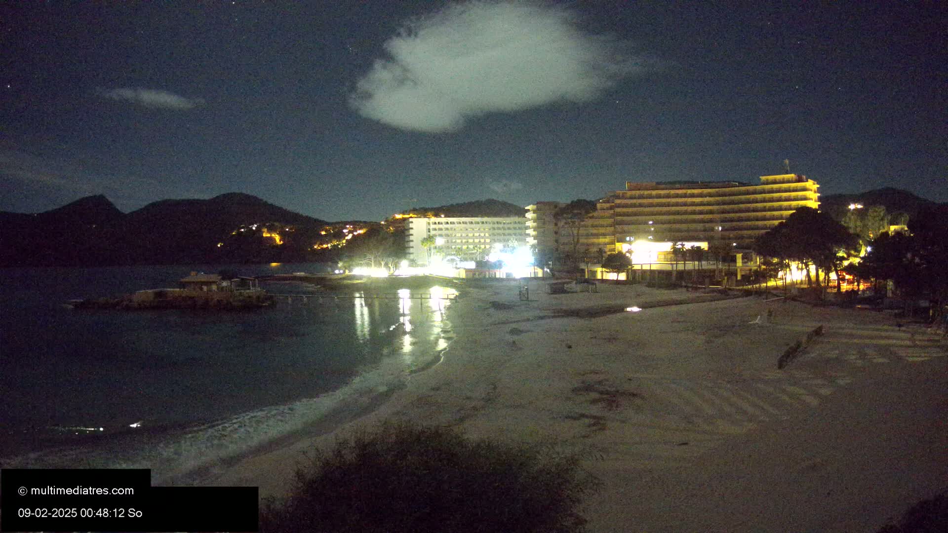 A nighttime beach scene shows a sandy shore, lit-up hotels, and calm water lapping against the sand under a partly cloudy sky.