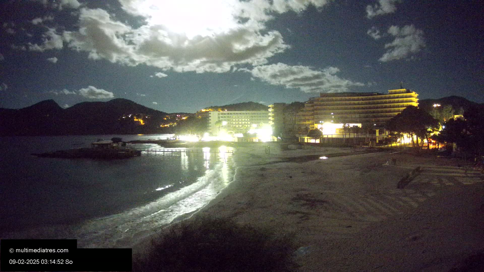 A moonlit night scene shows a sandy beach next to a calm body of water, with illuminated hotels and palm trees behind it, all under a partly cloudy sky.