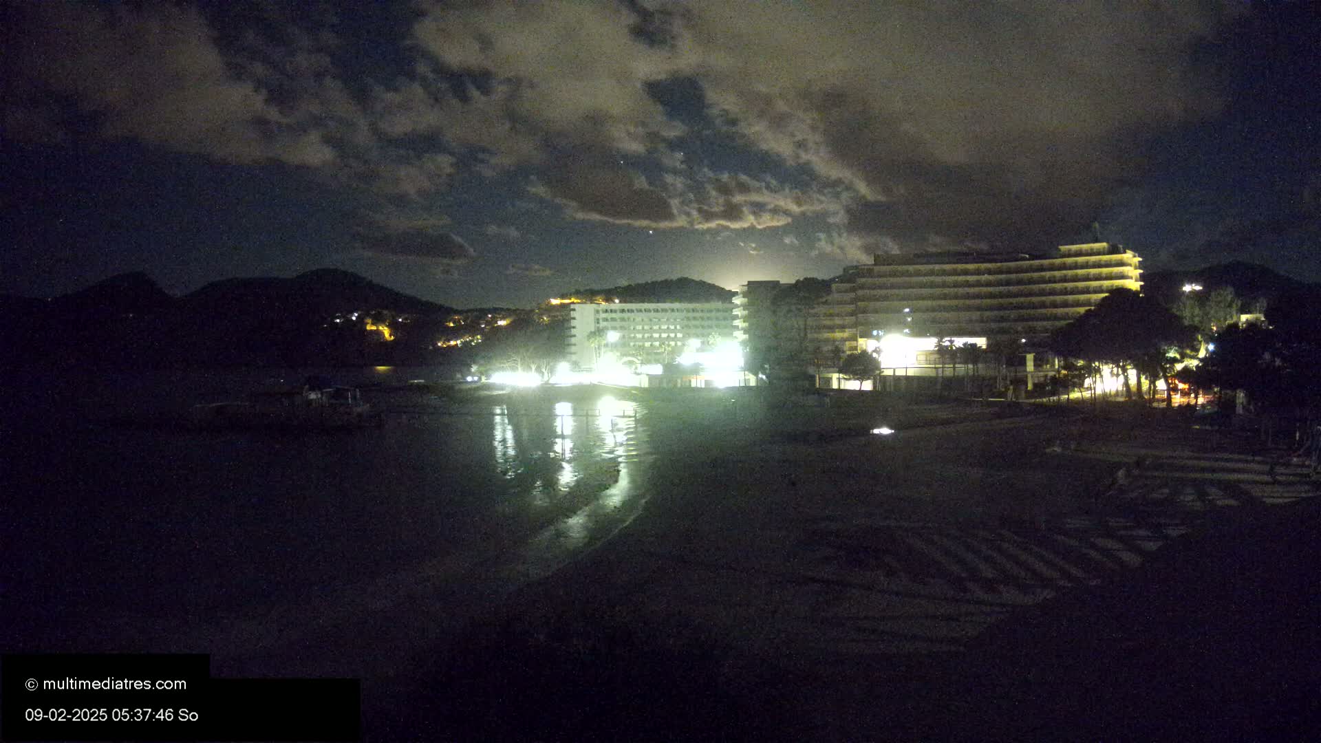 A nighttime view of a beach with two illuminated hotels and dark hills in the background under a cloudy sky.