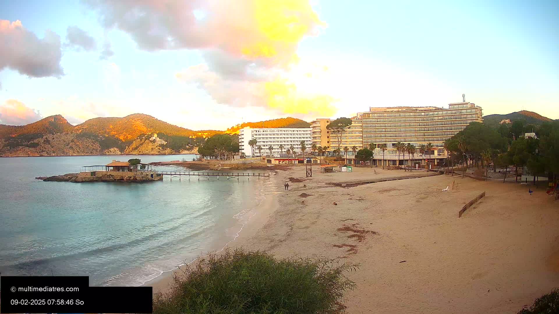 A sandy beach with calm, light blue water laps gently at the shore, backed by hotels and a small island with a structure, all under a partly cloudy, sunrise sky.