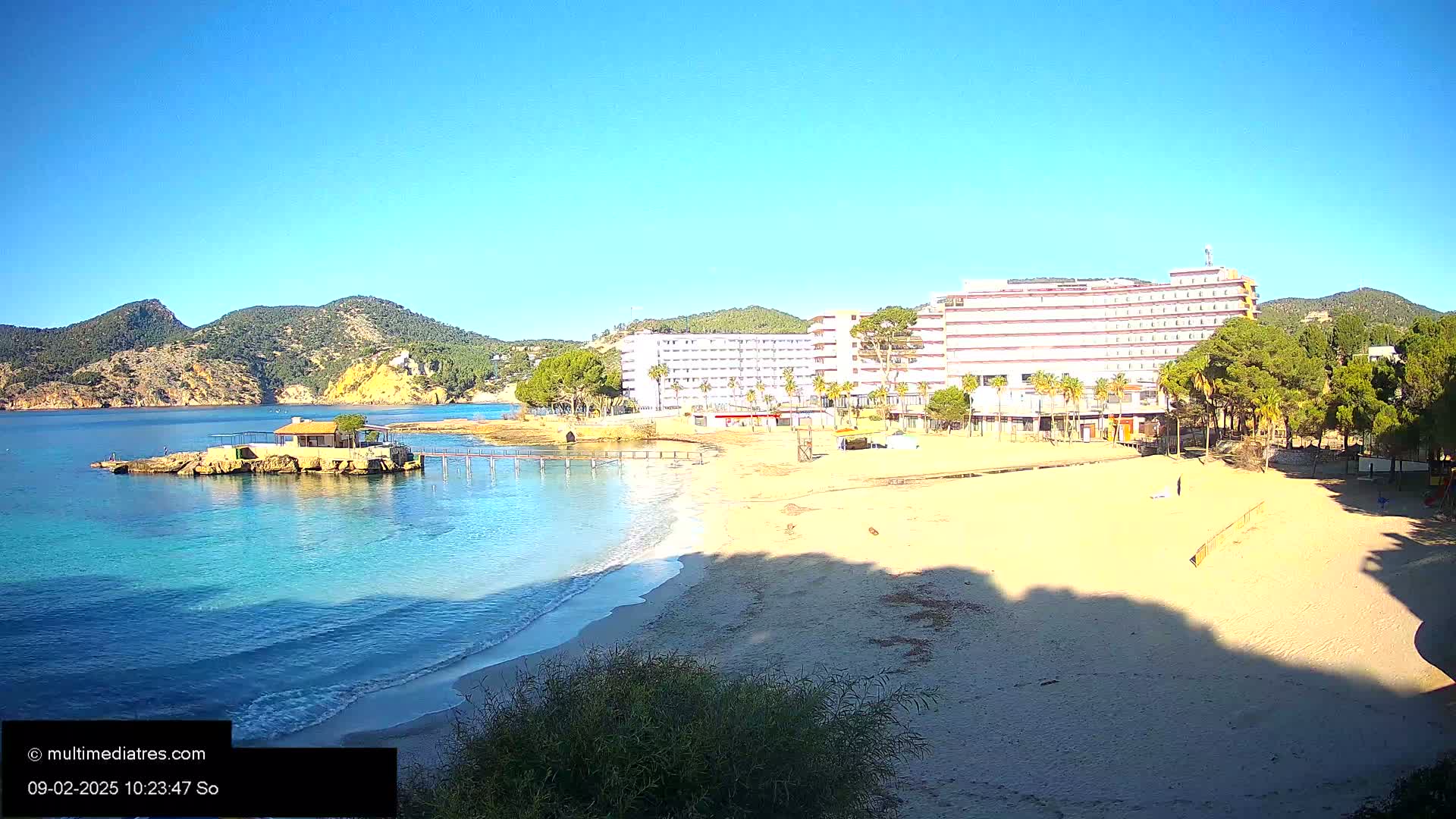 A sandy beach with gentle waves laps the shore near a hotel and a small building on a rock outcropping, all under a clear blue sky.