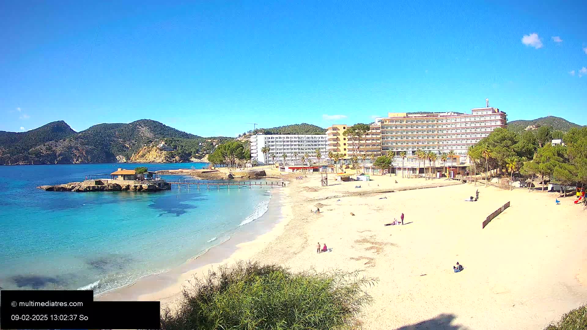 A sandy beach with turquoise water and several hotels sits under a clear blue sky near a mountainous coastline.