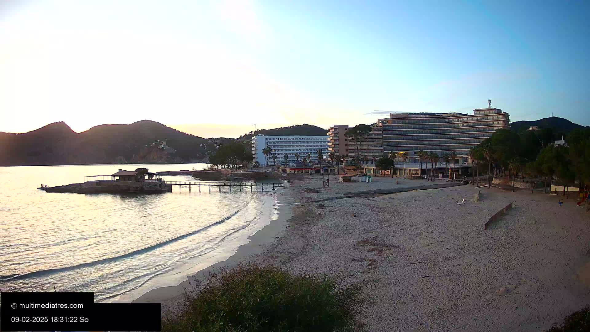 A sandy beach at sunset, with calm waves lapping the shore, overlooks a calm bay and several hotels nestled against a mountain.