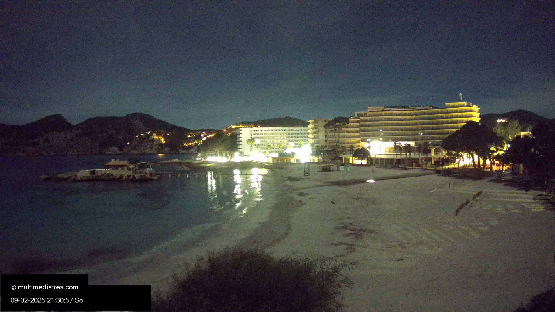 At night, a sandy beach is illuminated, with two lit hotels and a small building on a rocky outcropping in the water, all under a clear, dark sky, backed by dark mountains.