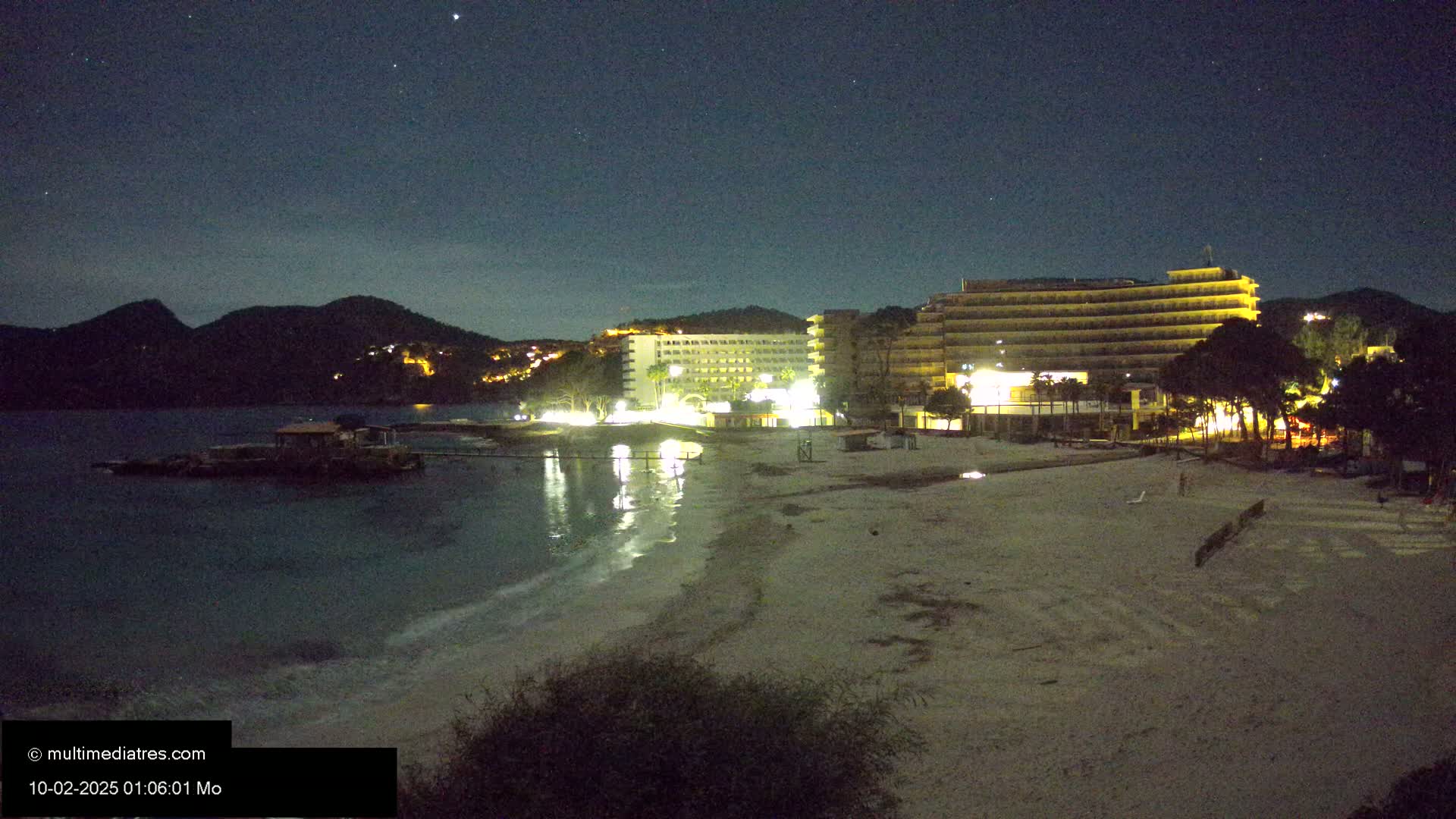 A nighttime view of a sandy beach with illuminated hotels and a calm sea under a clear, starry sky.