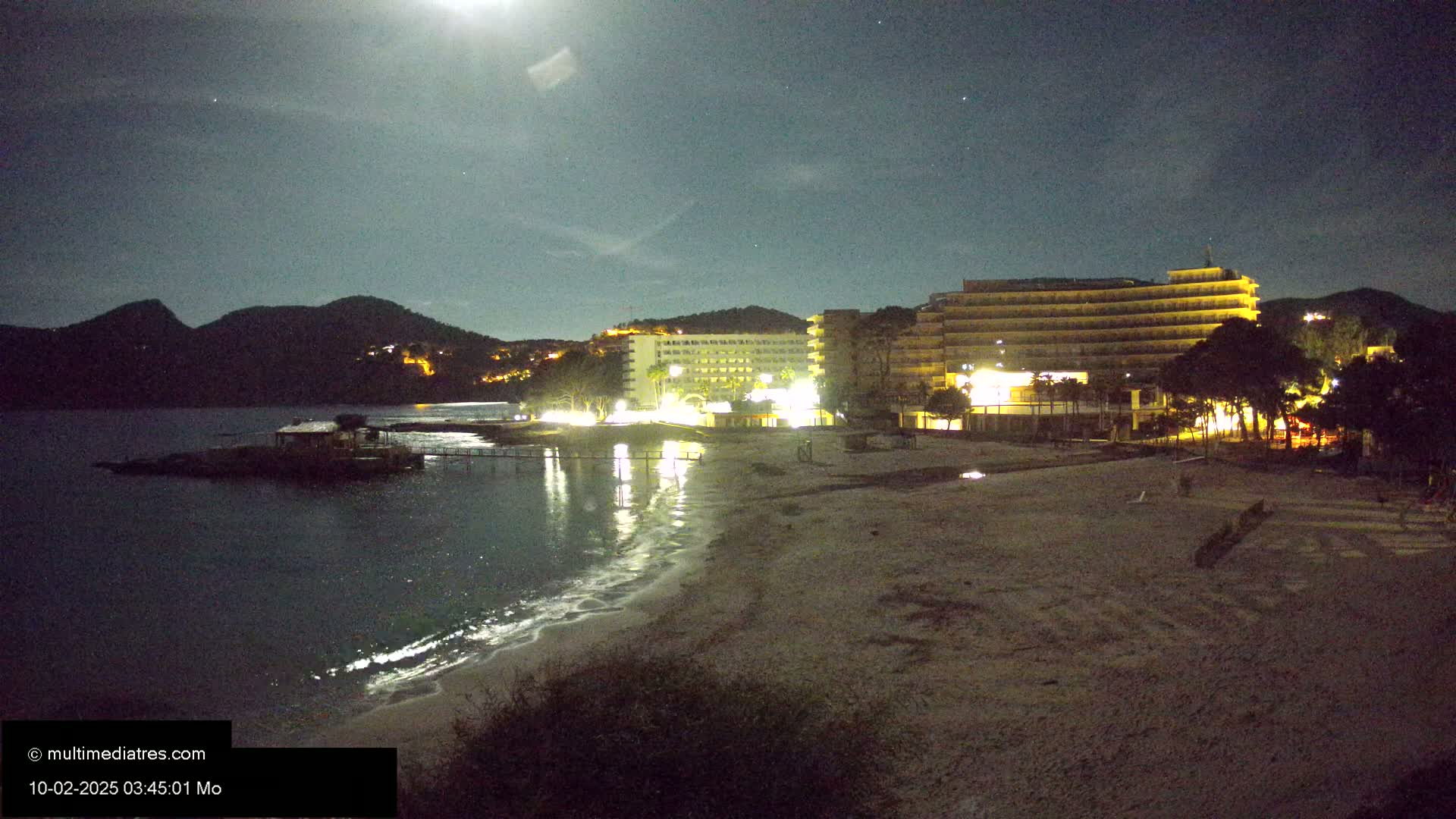 A moonlit beach at night shows a sandy shore, calm water, and illuminated hotels nestled against dark hills.