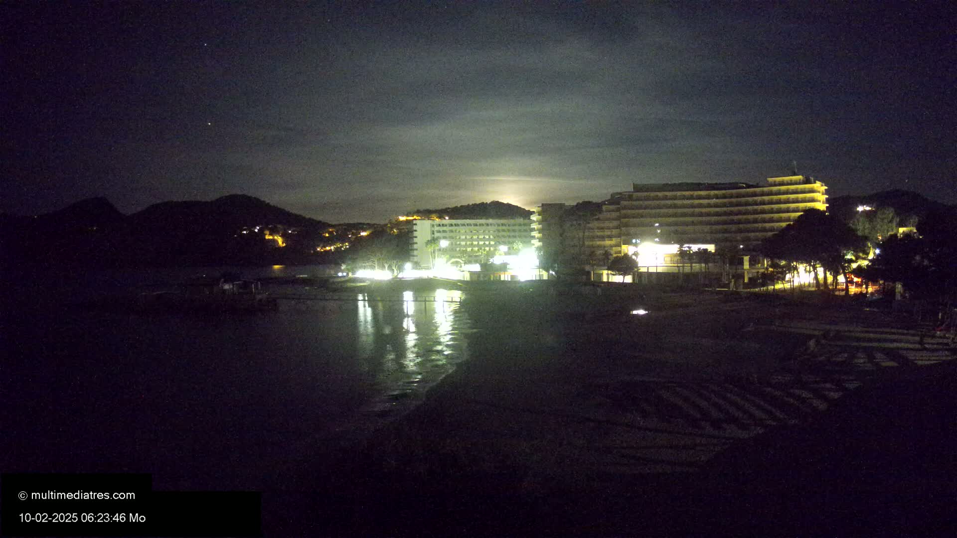 At night, under a partly cloudy sky, illuminated hotels sit on a beach next to a dark body of water and a hill.
