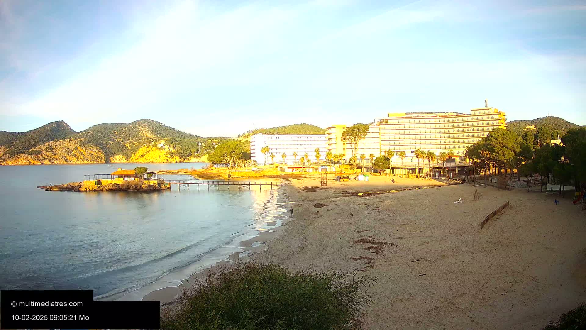 A sandy beach with calm, clear water laps at the shore near a large hotel, with hills and vegetation in the background under a mostly clear sky.