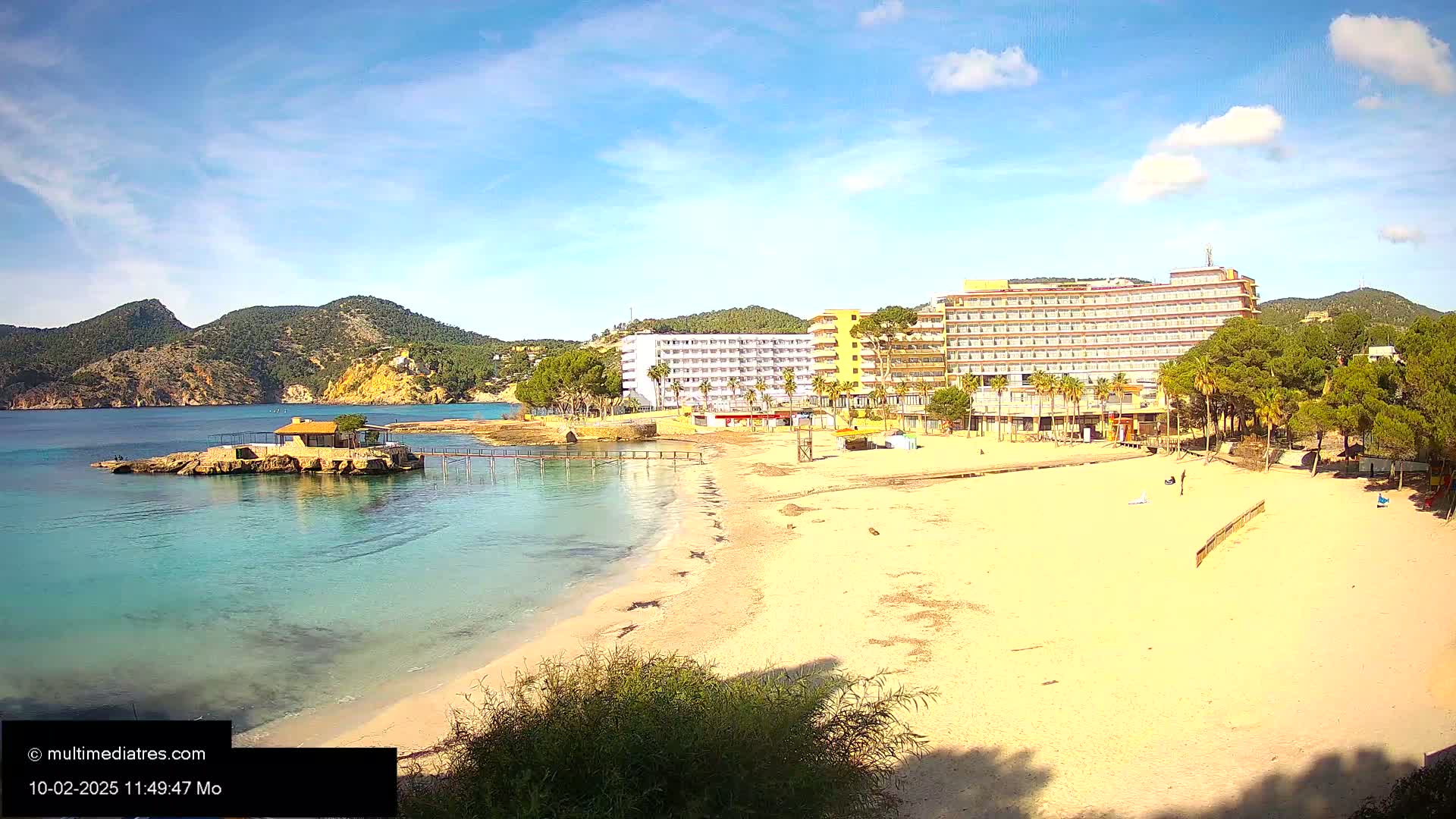 A sandy beach with calm, clear turquoise water, a small island with a building, and large hotels behind it under a mostly sunny sky.