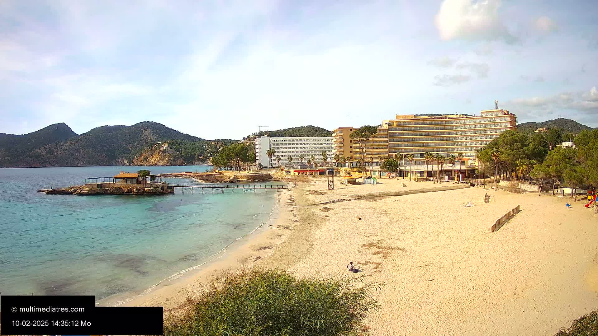 A sandy beach with a small building on a rock in the water, a pier, and large hotels behind it, all under a partly cloudy sky.