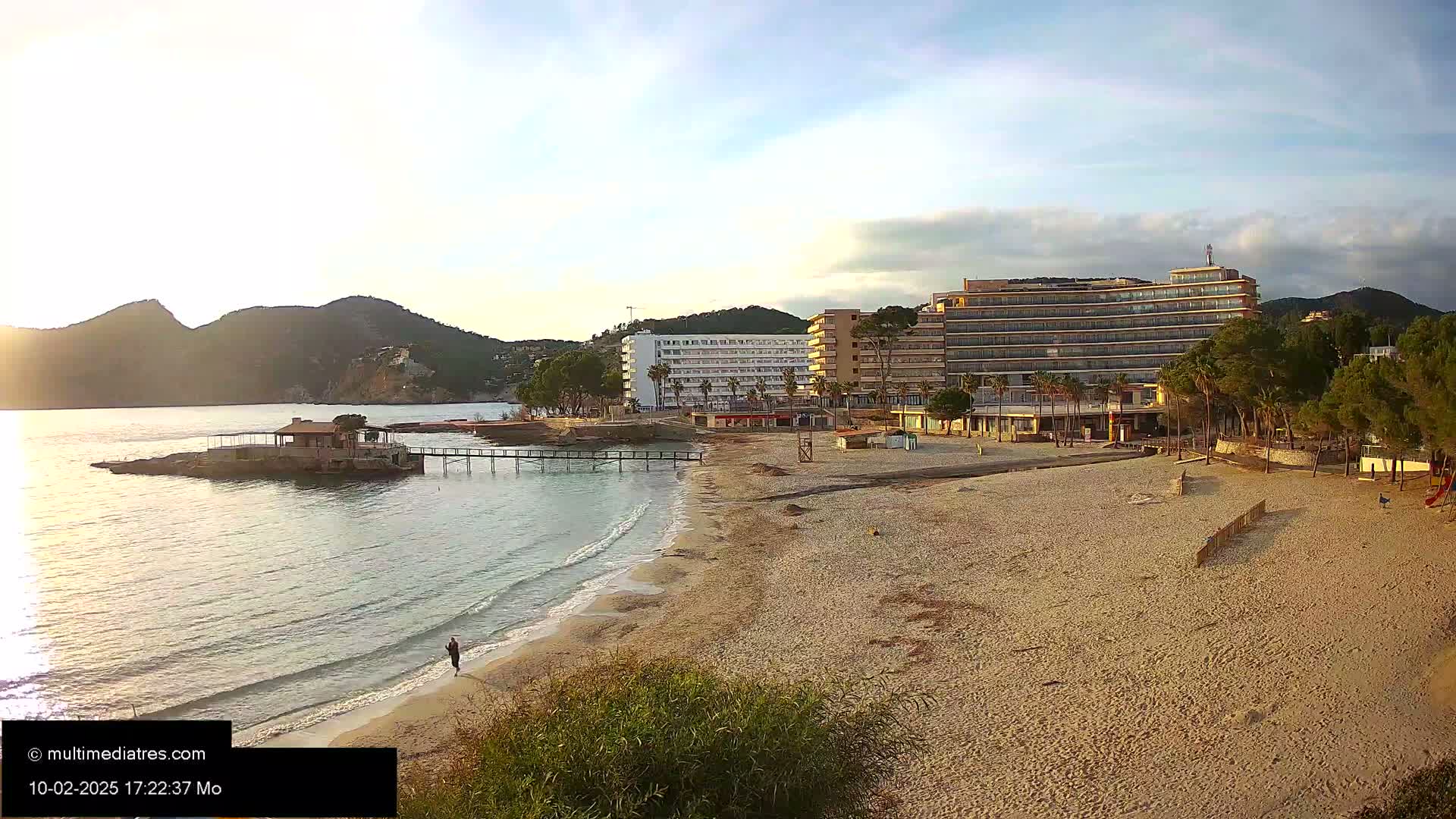 A sandy beach with calm waves laps against the shore, near hotels and a small pier, under a mostly clear sky.