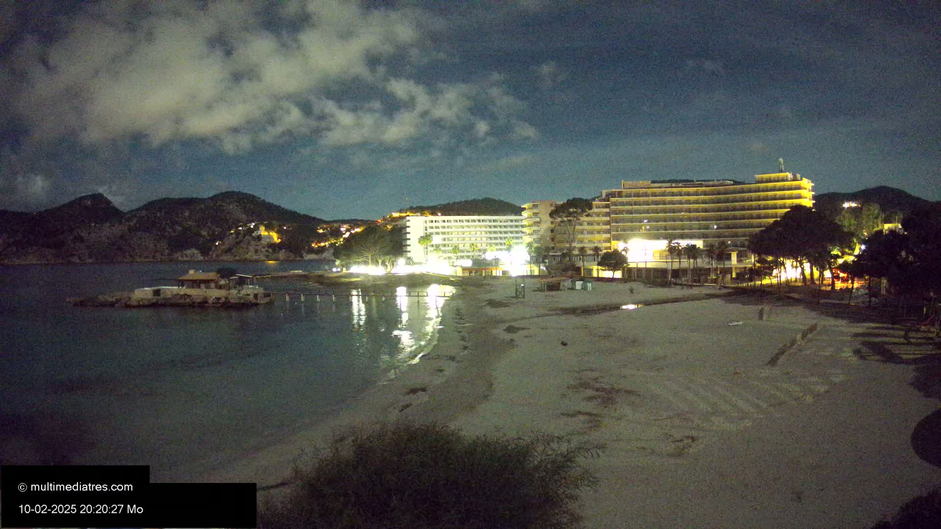 A nighttime beach scene shows two illuminated hotels behind a mostly empty sandy beach, with calm water and dark hills in the background under a cloudy sky.