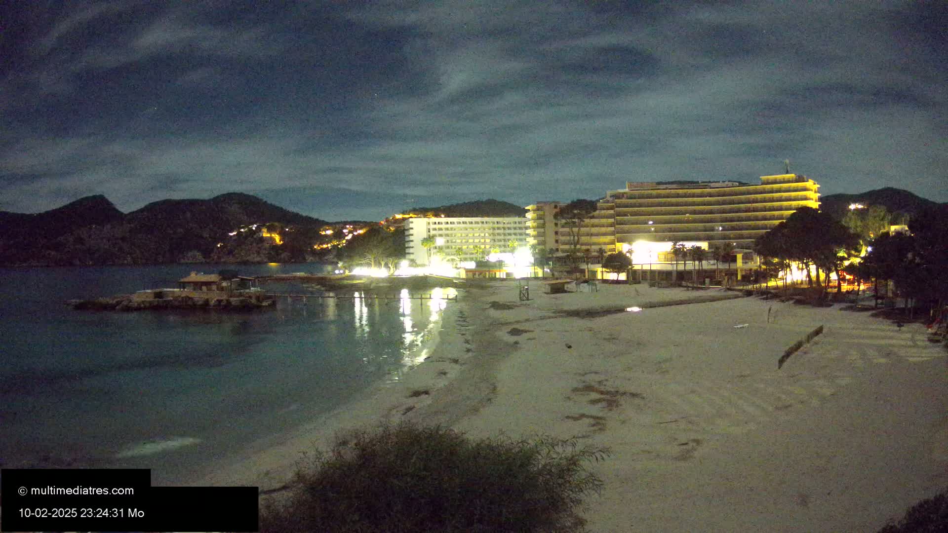 A nighttime view of a sandy beach with two illuminated hotels and a small island in a calm bay under a cloudy sky.