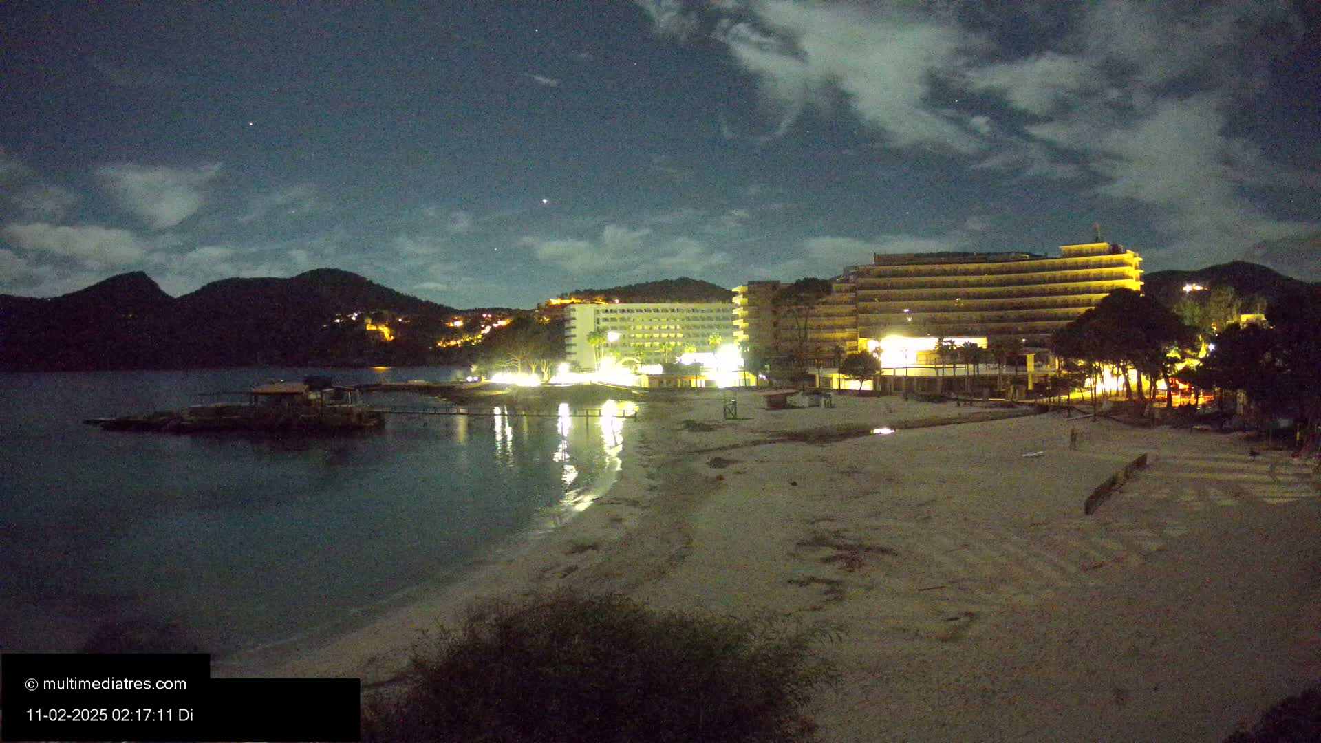 A moonlit beach at night with two illuminated hotels and a calm body of water, backed by dark hills under a partly cloudy sky.