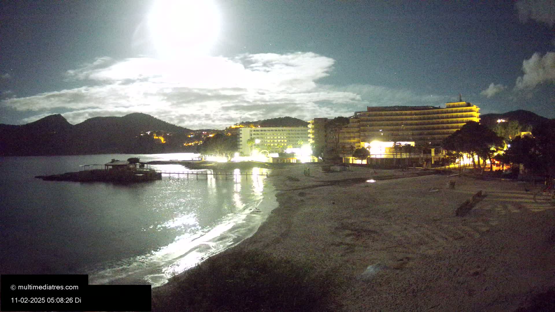 A moonlit night scene shows a sandy beach, calm water, and illuminated hotels near a mountainous coastline.