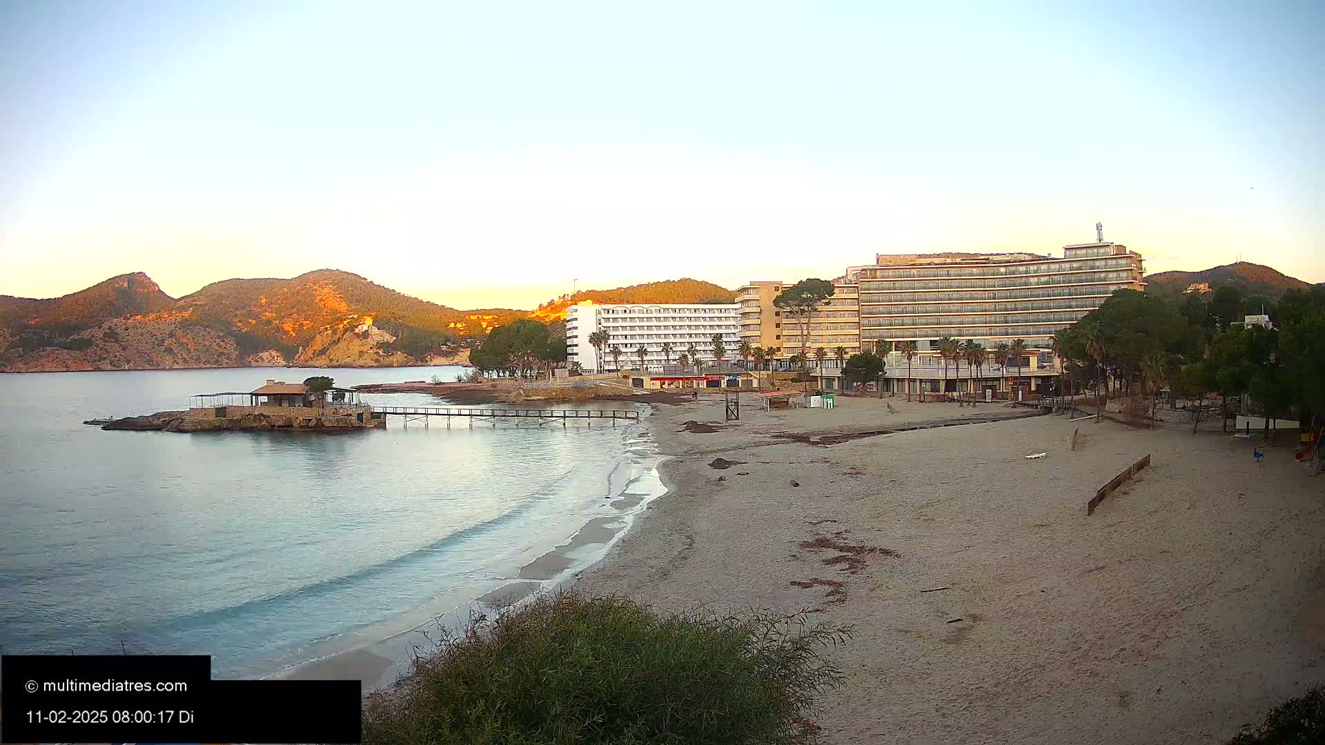 A sandy beach with calm, clear water and two hotels sits near a rocky shoreline and mountains under a clear, sunrise sky.