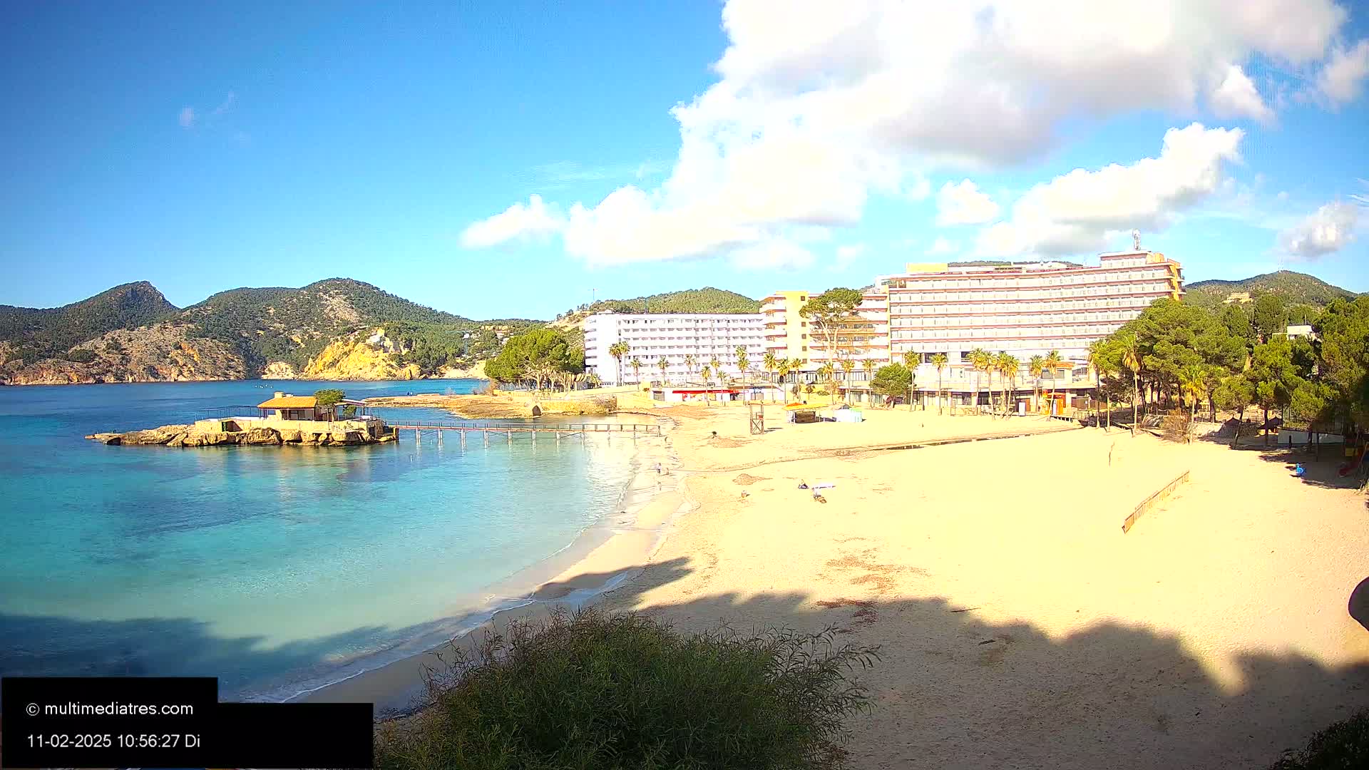 A sandy beach with a small, calm bay, a hotel, and a small building on a rock outcropping under a partly cloudy, sunny sky.