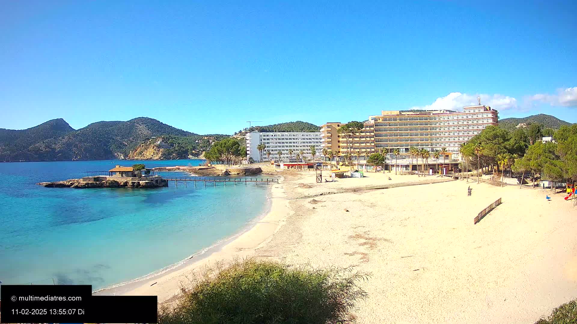 A sandy beach with turquoise water, a small island with a building, and a large hotel in the background under a clear, sunny sky.
