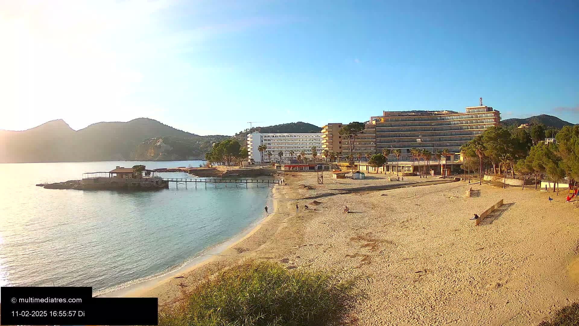 A sandy beach with a few people on it stretches out in front of several hotels and a line of trees under a sunny sky, with mountains visible in the distance across calm water.