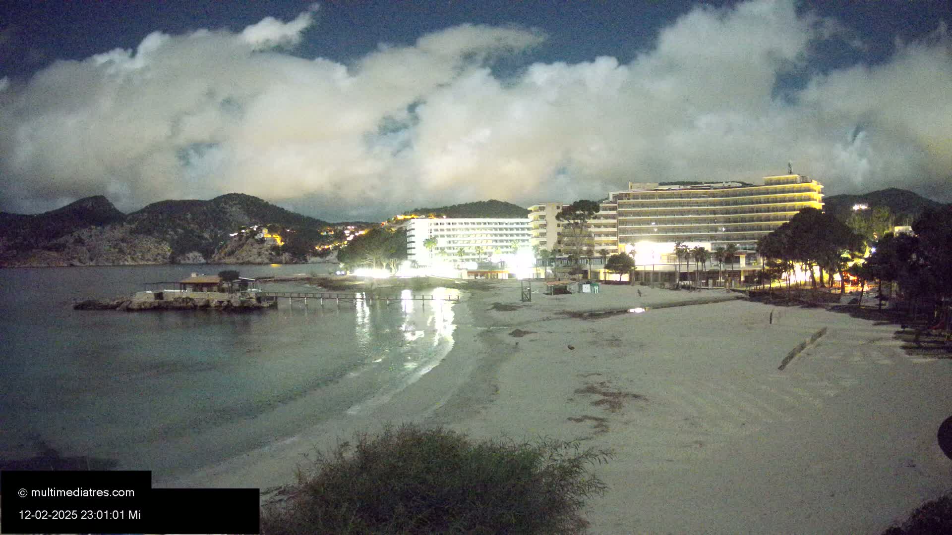 A nighttime beach scene shows two illuminated hotels near a calm, dark-blue sea under a cloudy sky.