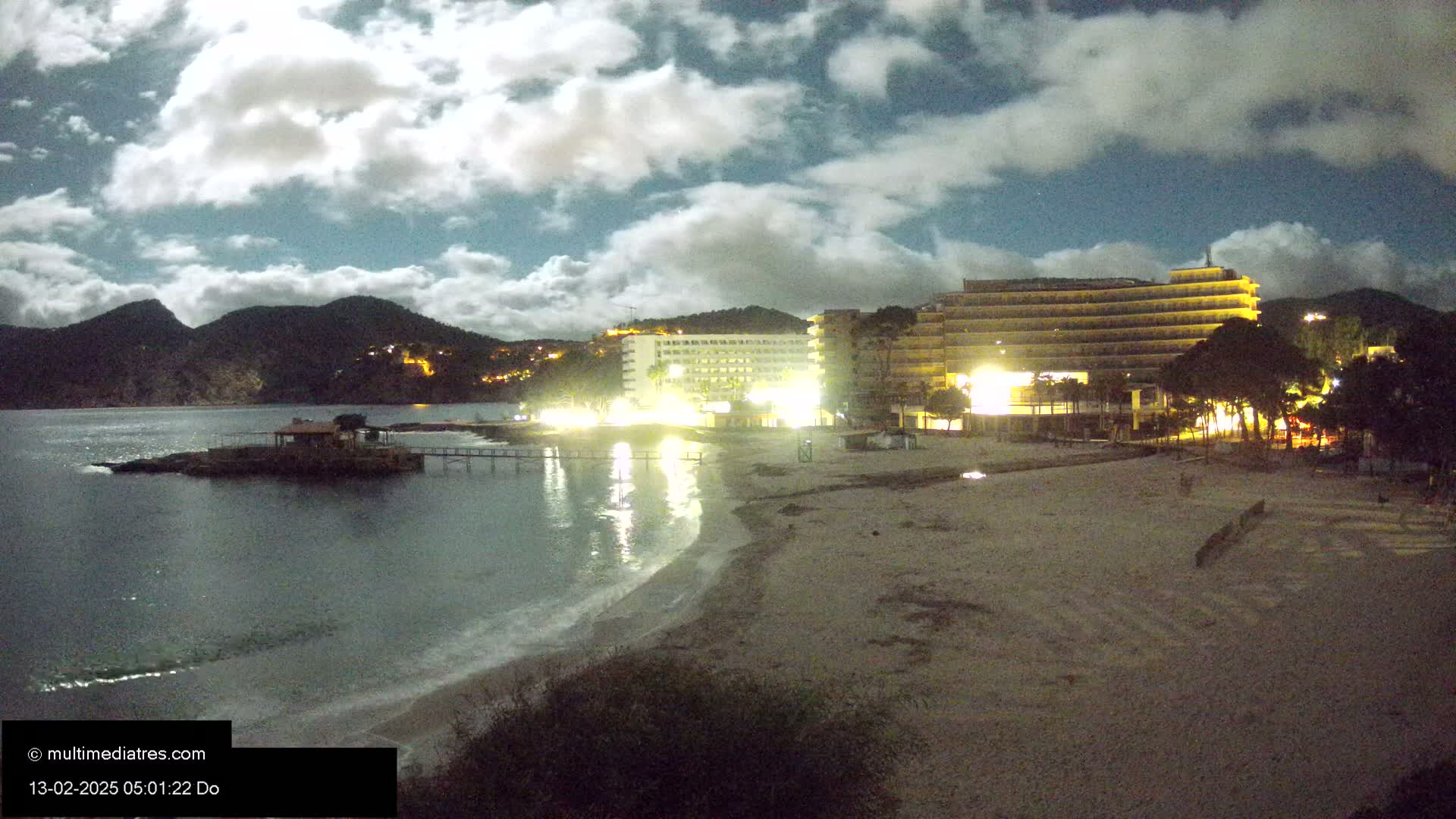 A moonlit beach at night with gently lapping waves,  illuminated hotels, and dark hills in the background under a partly cloudy sky.