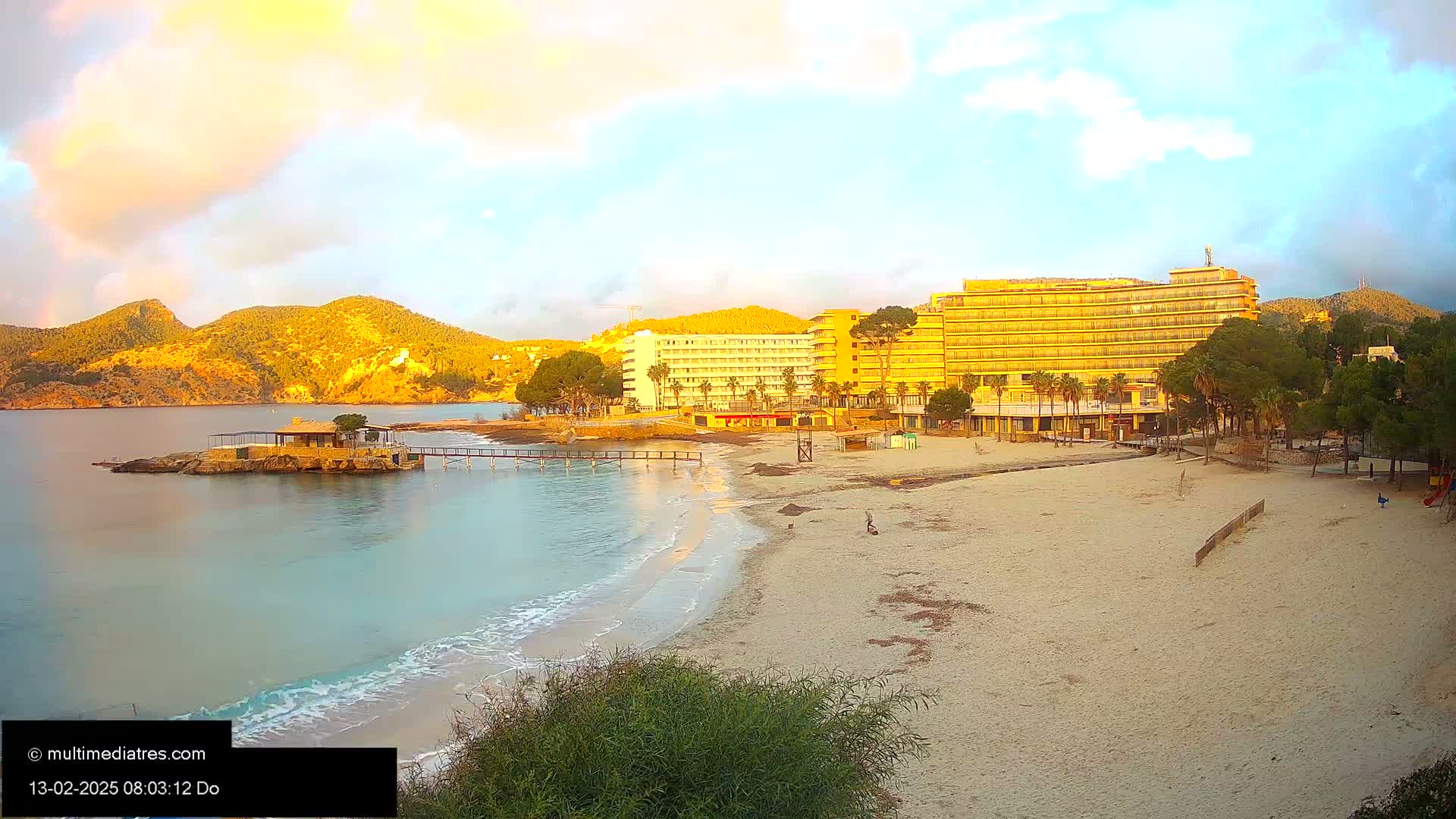 A sandy beach with calm, turquoise water laps gently at the shore, under a partly cloudy, golden sunrise; behind the beach are several hotels and green hills.