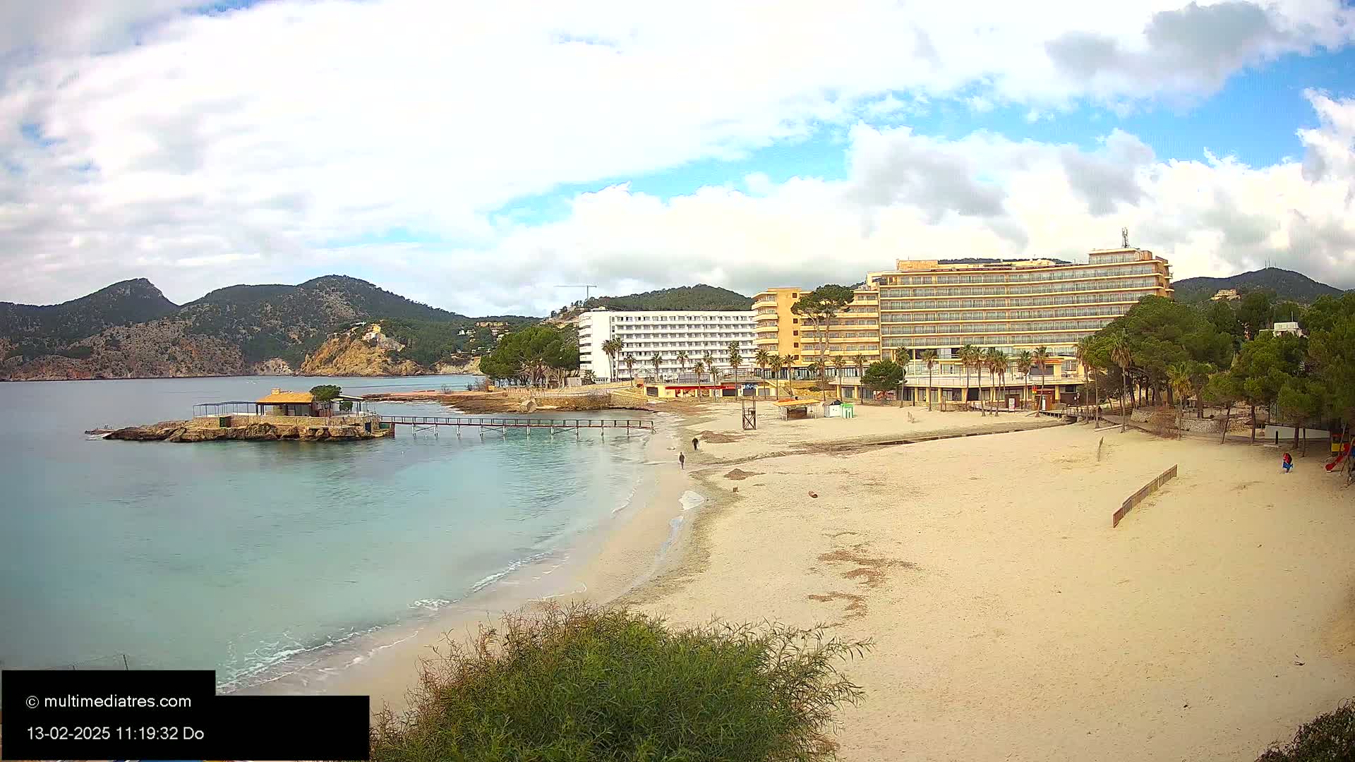 A sandy beach with calm, light-blue water and two hotels behind it, under a partly cloudy sky.