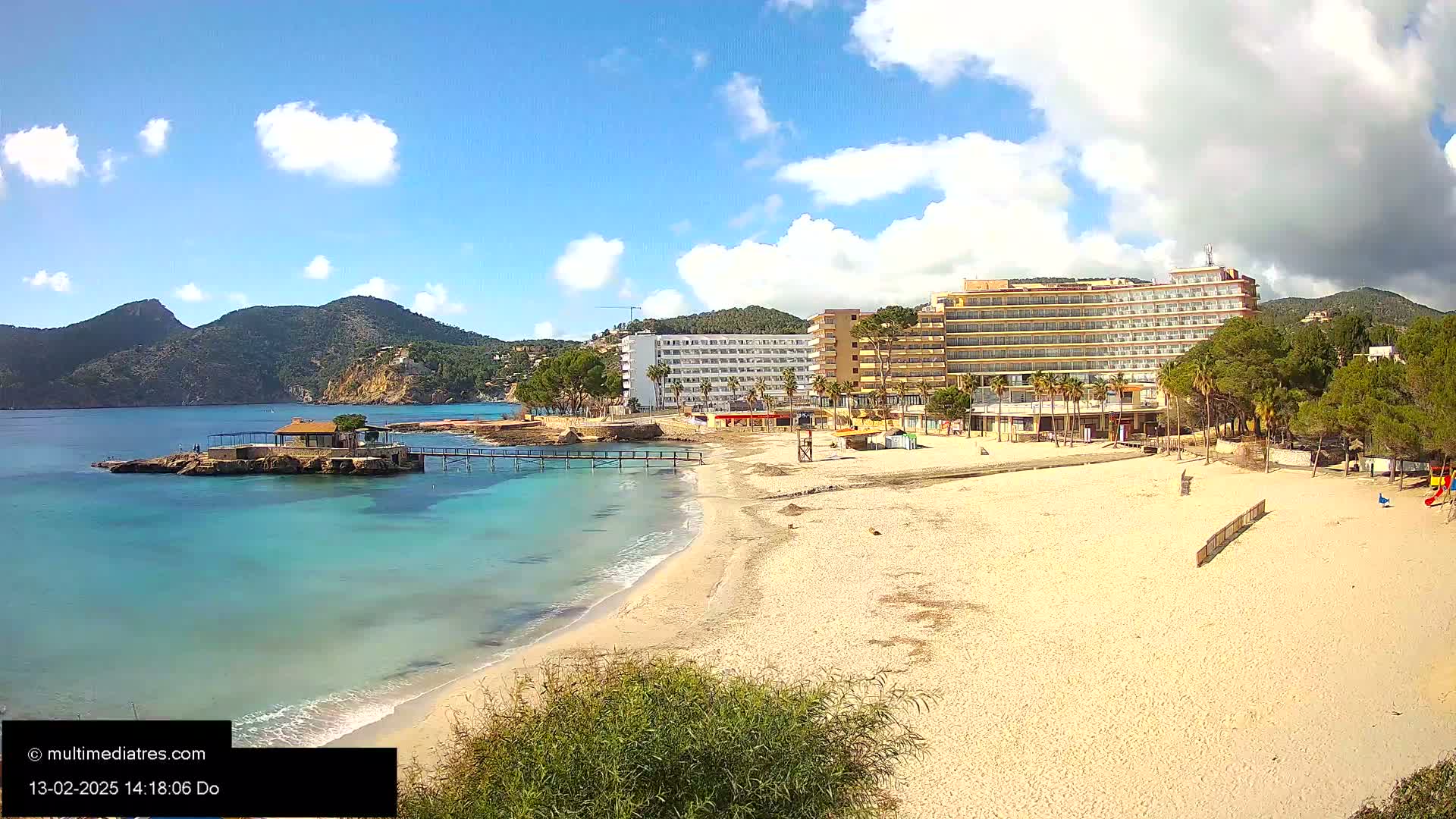 A sandy beach stretches toward a turquoise ocean, with a small island, a pier, and large hotels situated nearby under a partly cloudy sky.
