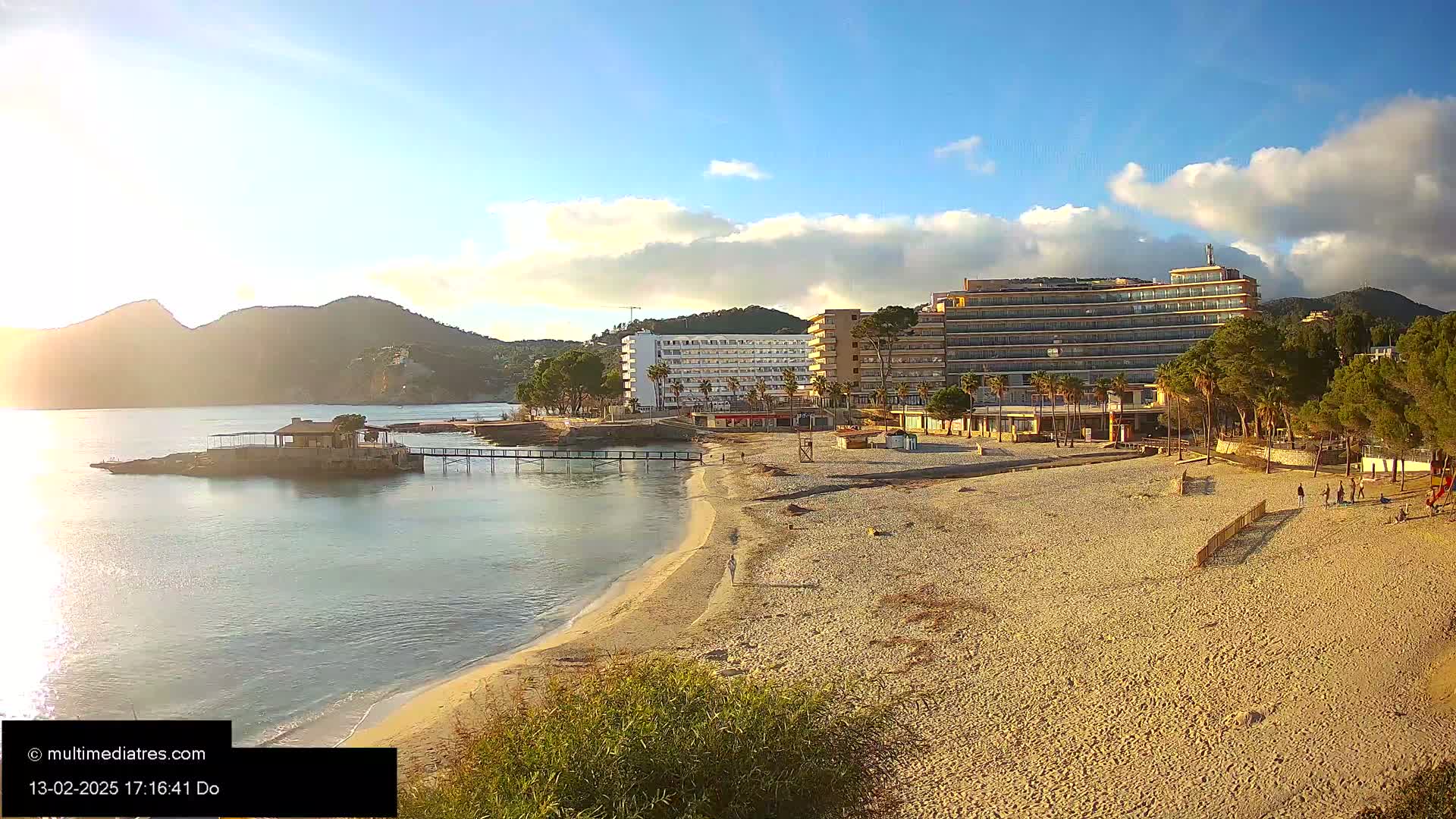 A sandy beach with several hotels and a small pier stretches along a calm bay under a partly cloudy sky.