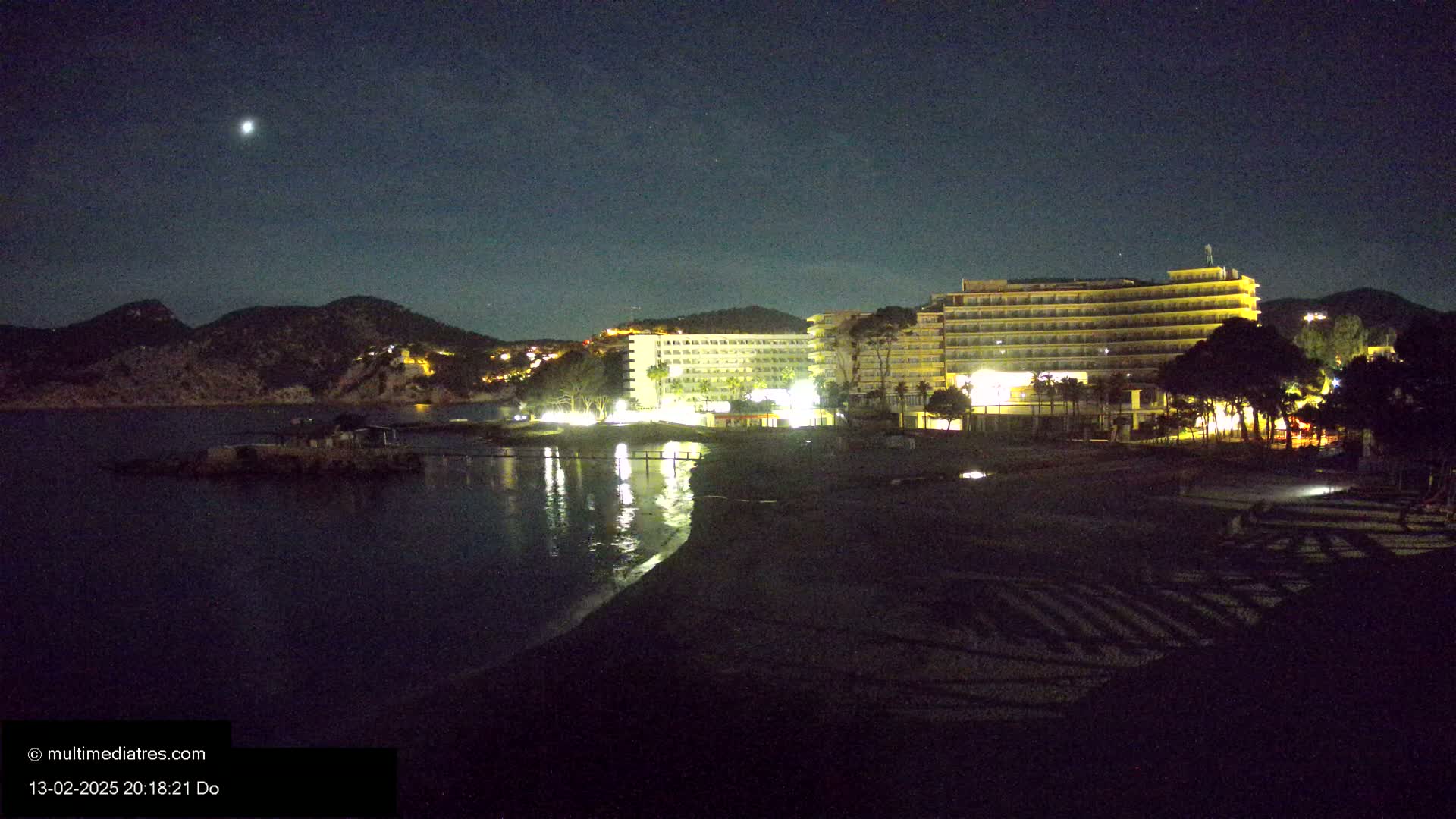 A dark beach at night, under a clear sky with a visible moon, is bordered by illuminated hotels and hills.