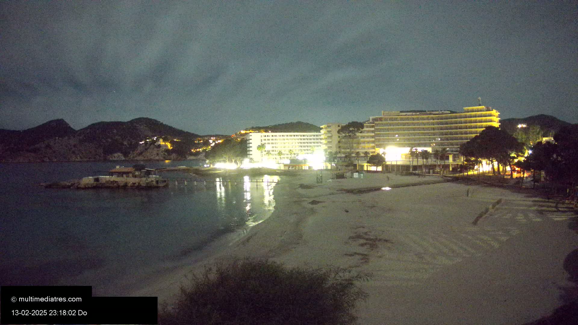 At night, a sandy beach is shown with illuminated hotels and a calm body of water under a dark, cloudy sky.