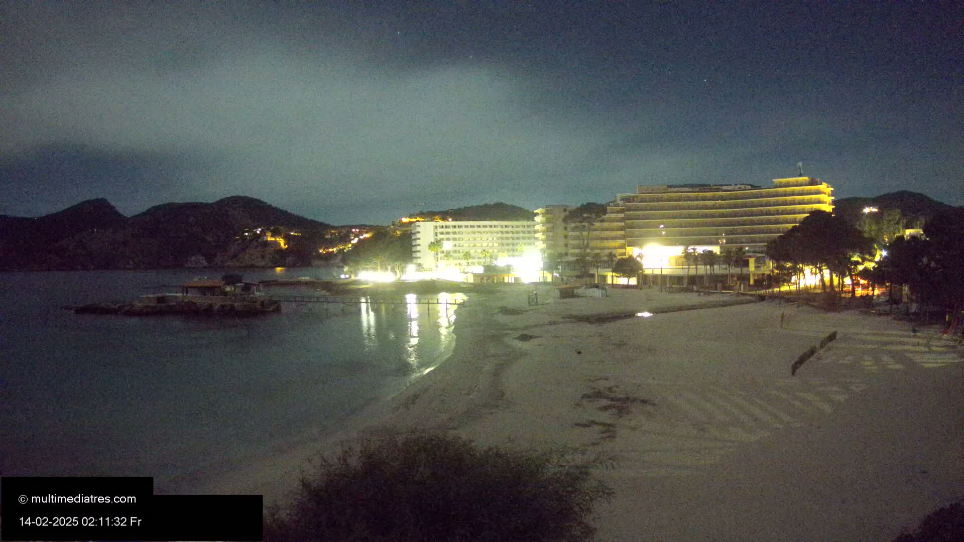 At night, under a mostly cloudy sky, a sandy beach is illuminated, with two large hotels and some trees visible beyond it.