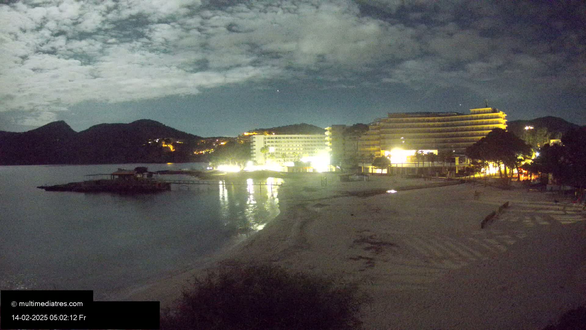 A moonlit beach at night shows two brightly lit hotels near the water's edge, with dark mountains in the background under a cloudy sky.