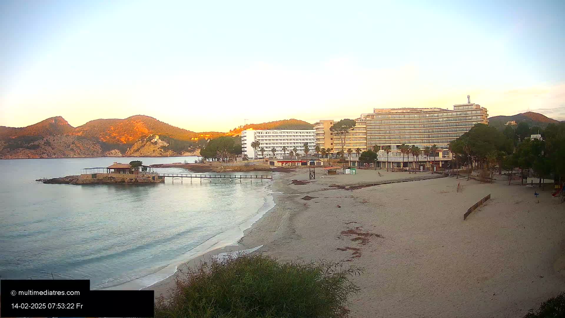 A sandy beach stretches toward two hotels situated near a calm bay, backed by sunlit hills under a clear morning sky.