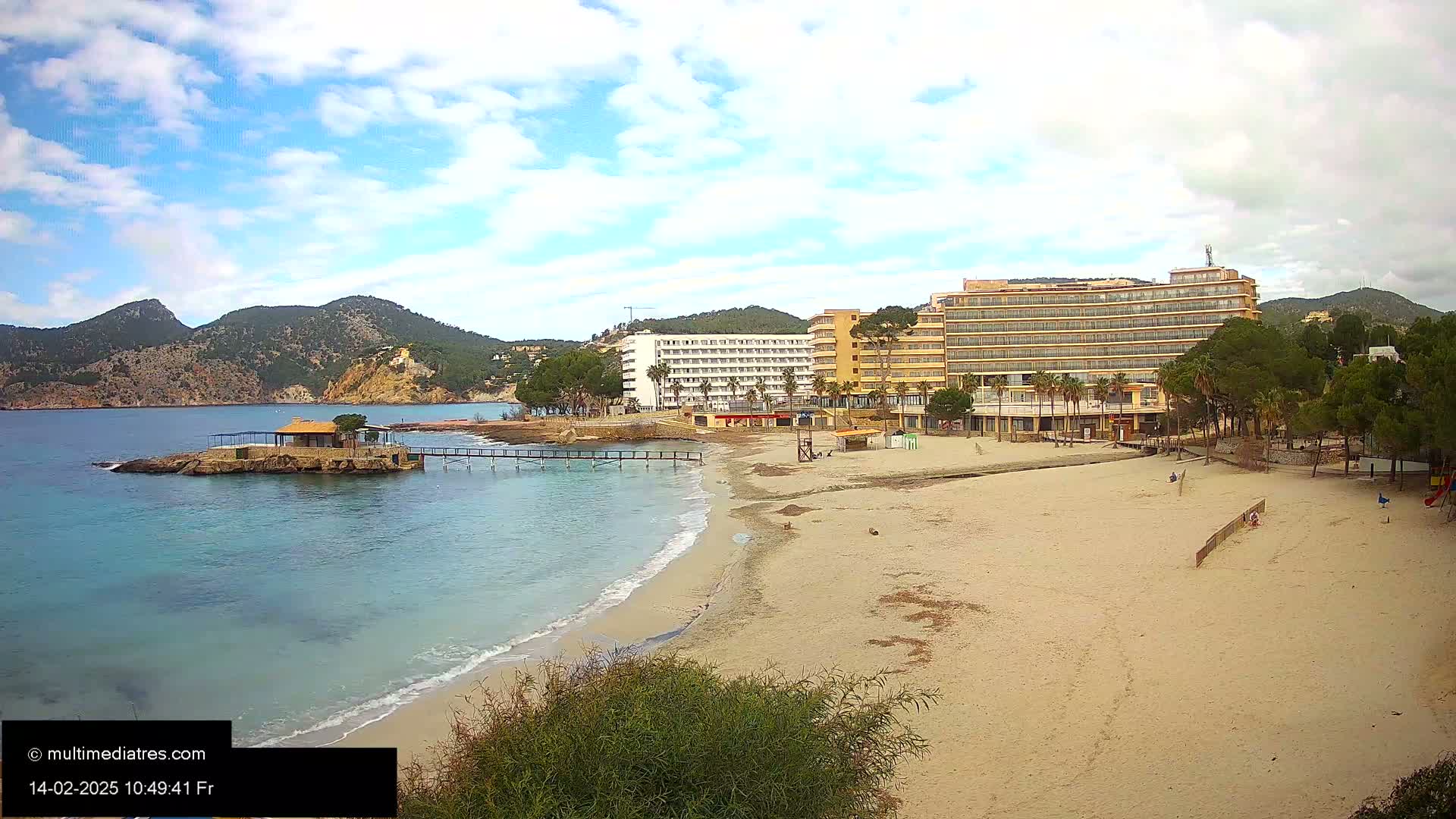 A sandy beach with calm, turquoise water, backed by hotels and green hills under a partly cloudy sky.