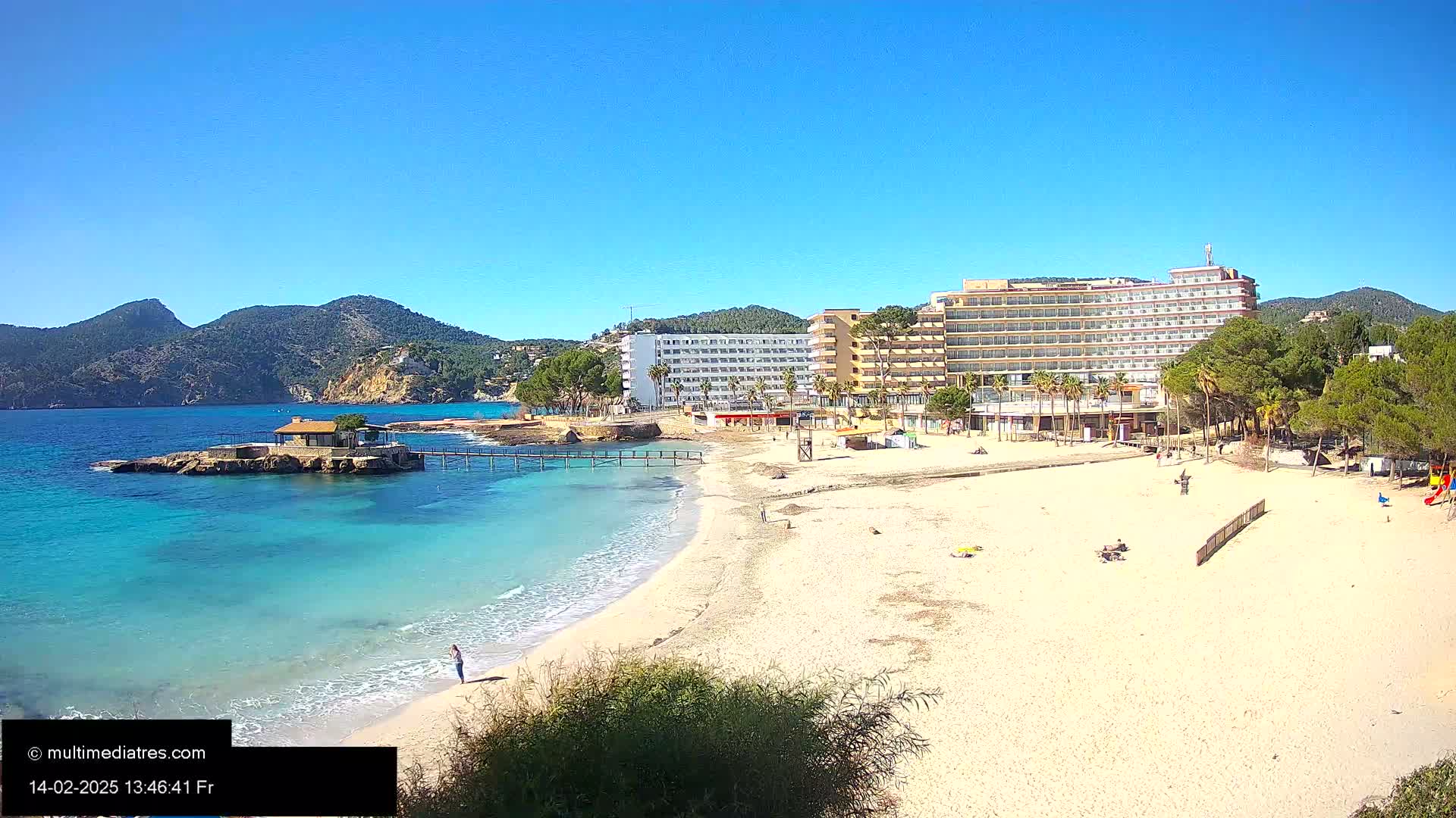 A sandy beach with calm, turquoise water laps the shore near a hotel complex and mountains under a clear blue sky.