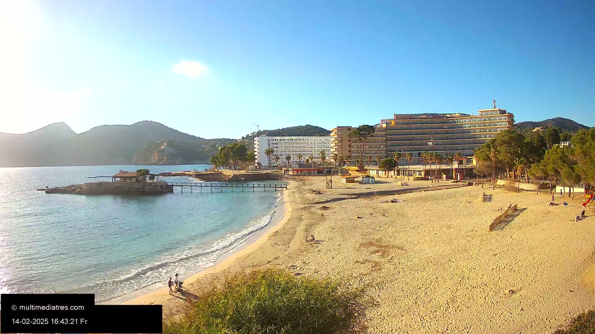A sandy beach stretches toward a calm sea next to a beachfront hotel and a small rocky island with a pier under a clear, sunny sky.