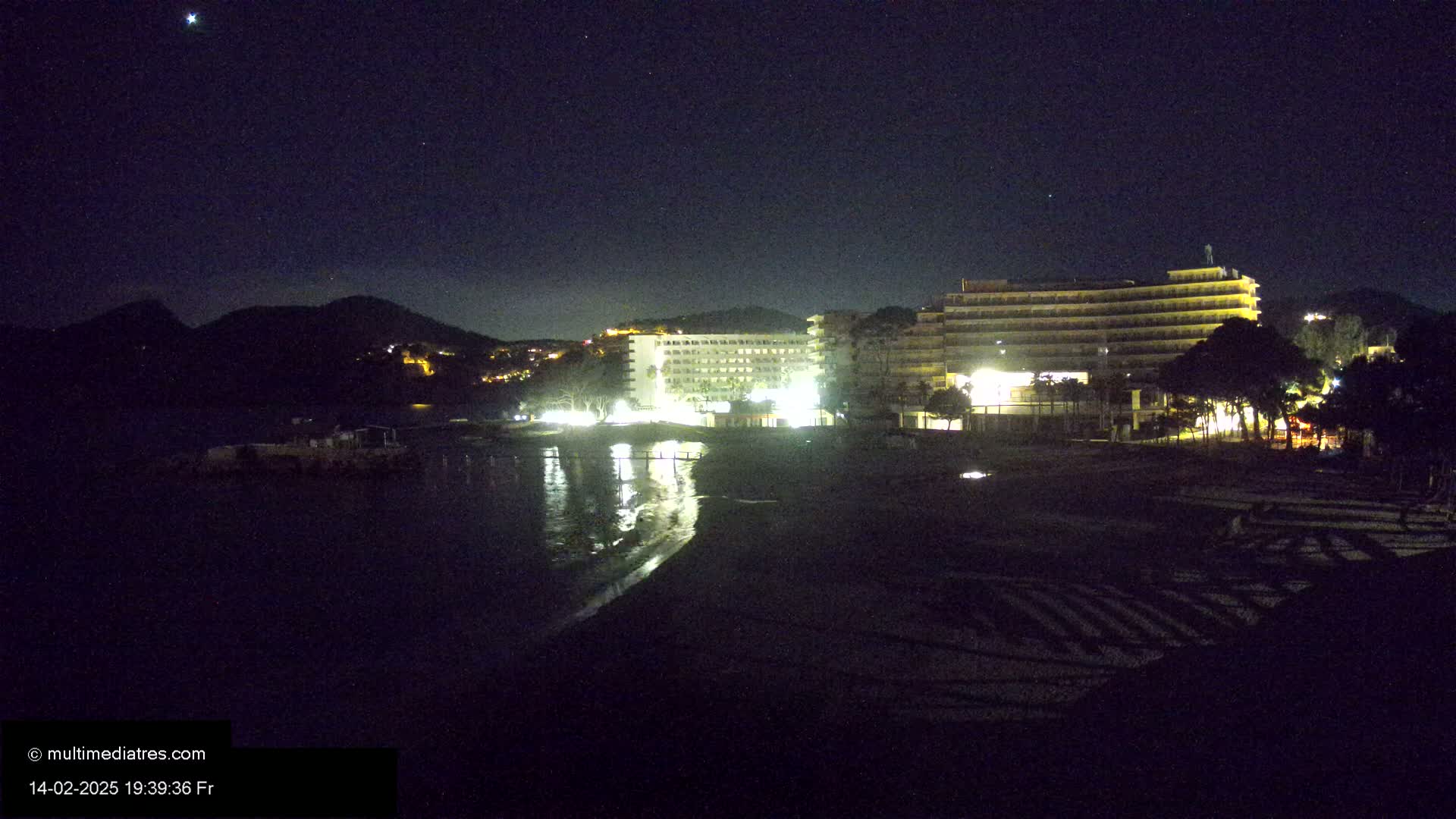 A nighttime view of a beach with two illuminated hotels and hills in the background under a clear, starry sky.