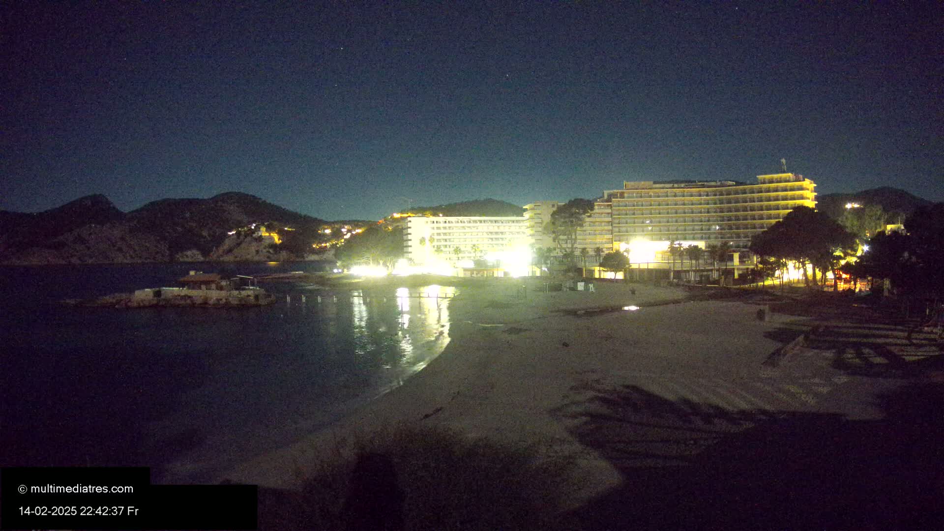A nighttime view of a beach with illuminated hotels and a calm sea under a clear, starry sky.