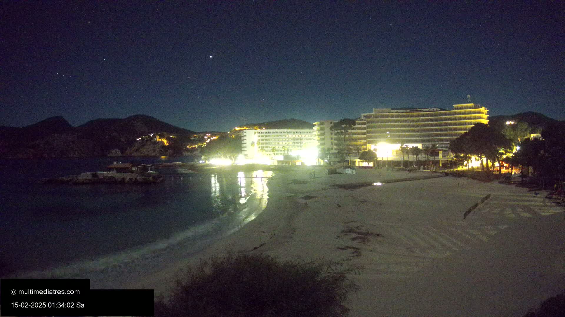 A sandy beach at night is illuminated by the lights of several hotels nestled against dark hills under a starry sky.