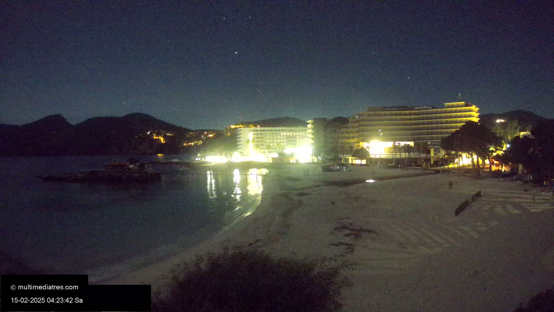 A nighttime beach scene shows a sandy shore, calm water reflecting the lights of nearby hotels and hills under a clear, starry sky.