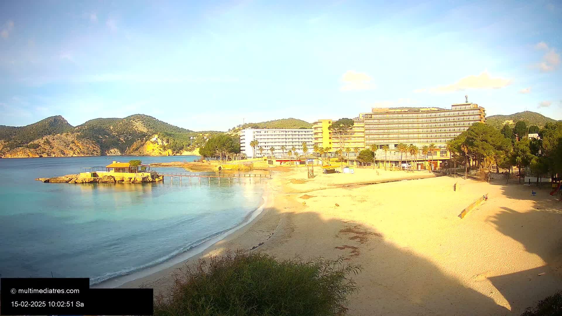 A sandy beach with calm, clear water laps at its shore, backed by several hotels and a small building on a rocky outcrop, all under a mostly sunny sky.