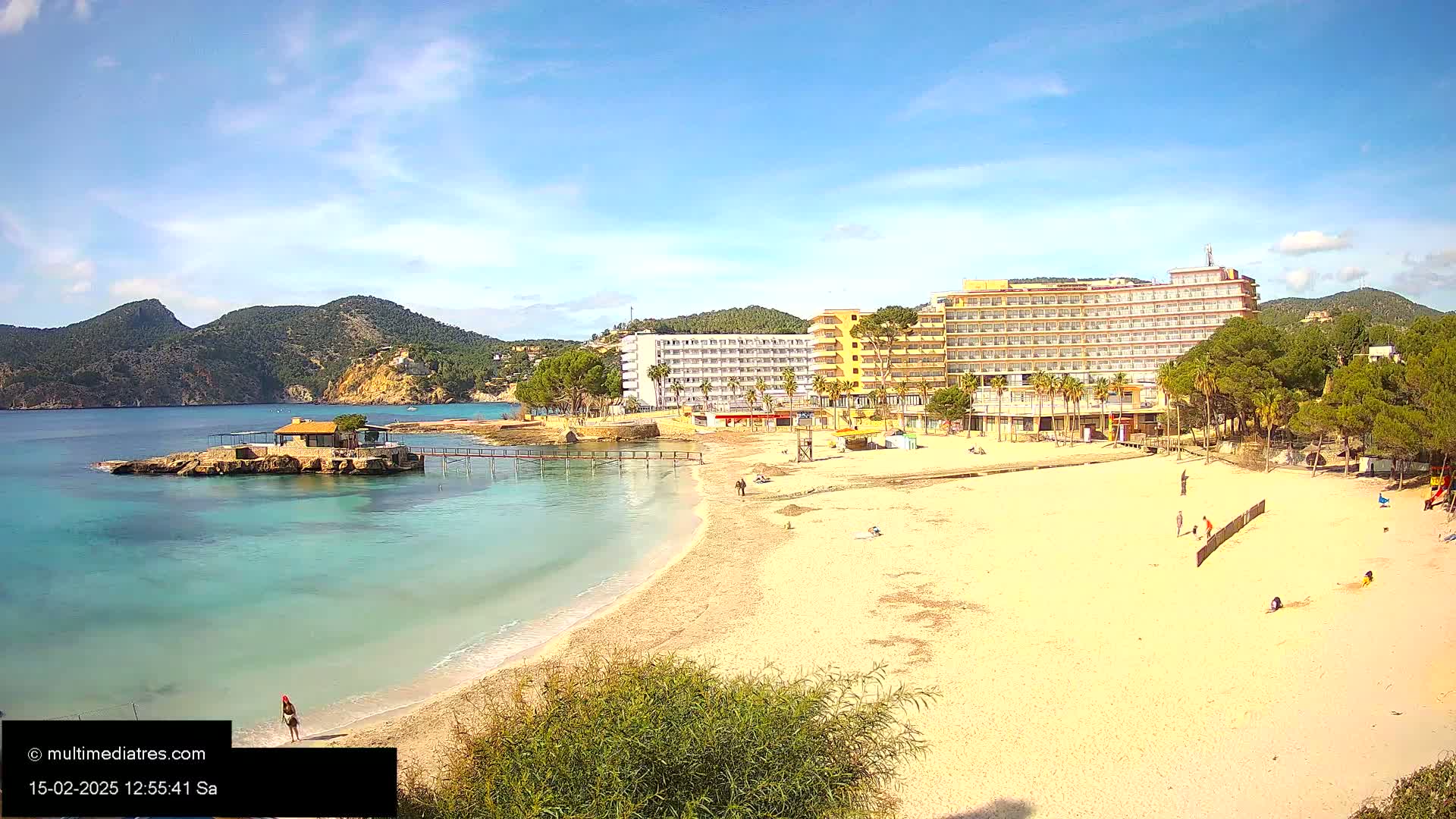 A sandy beach with calm, turquoise water, a small island with a building, and large hotels sits under a mostly sunny sky.