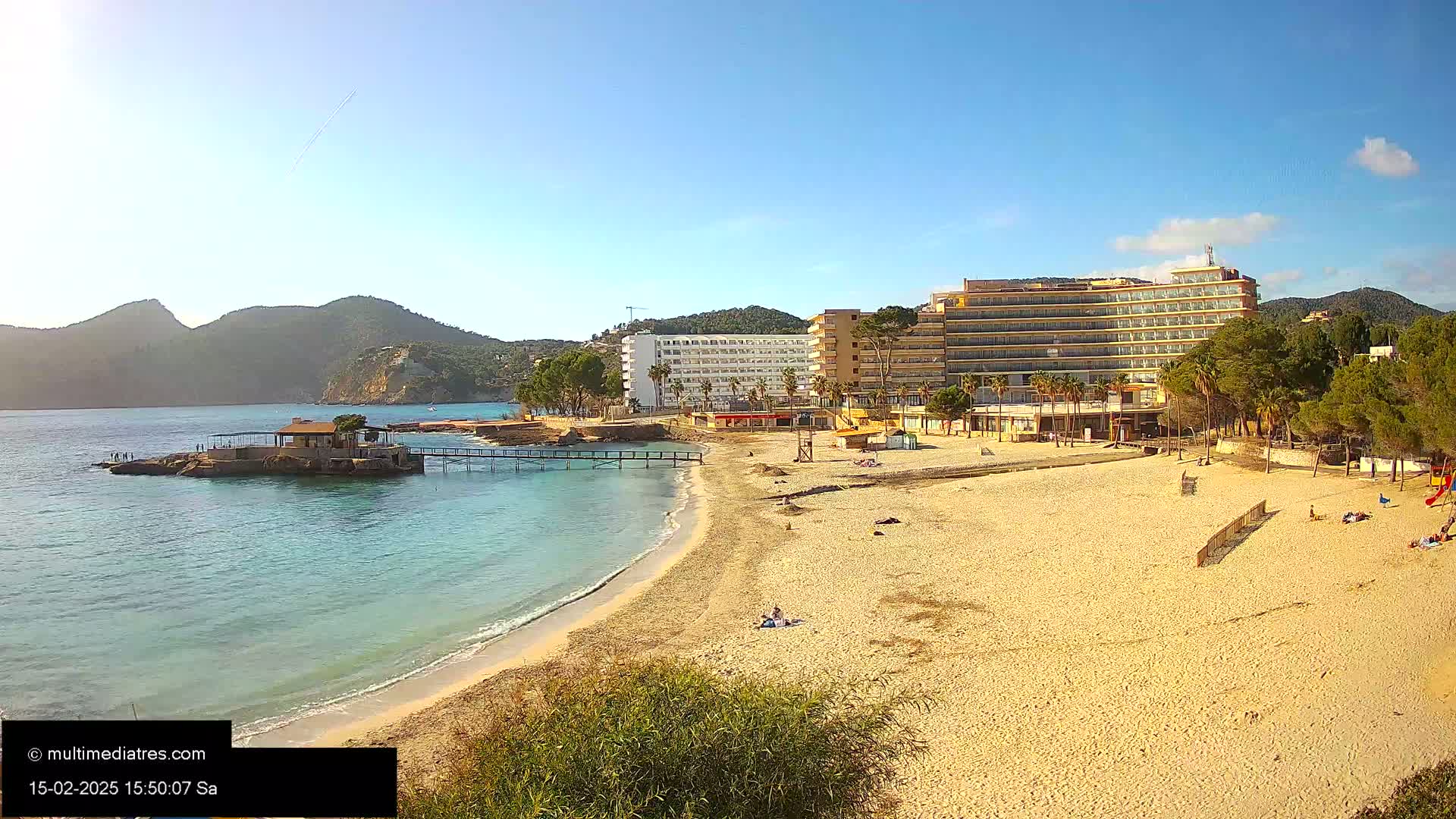 A sandy beach with a few people on it, a small pier, and several hotels behind it, all situated on a calm, clear bay near some mountains under a sunny sky.