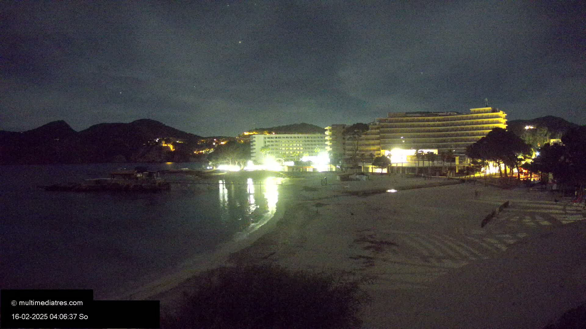 A nighttime view of a sandy beach and two illuminated hotels, with dark hills and a calm body of water in the background under a clear, starry sky.