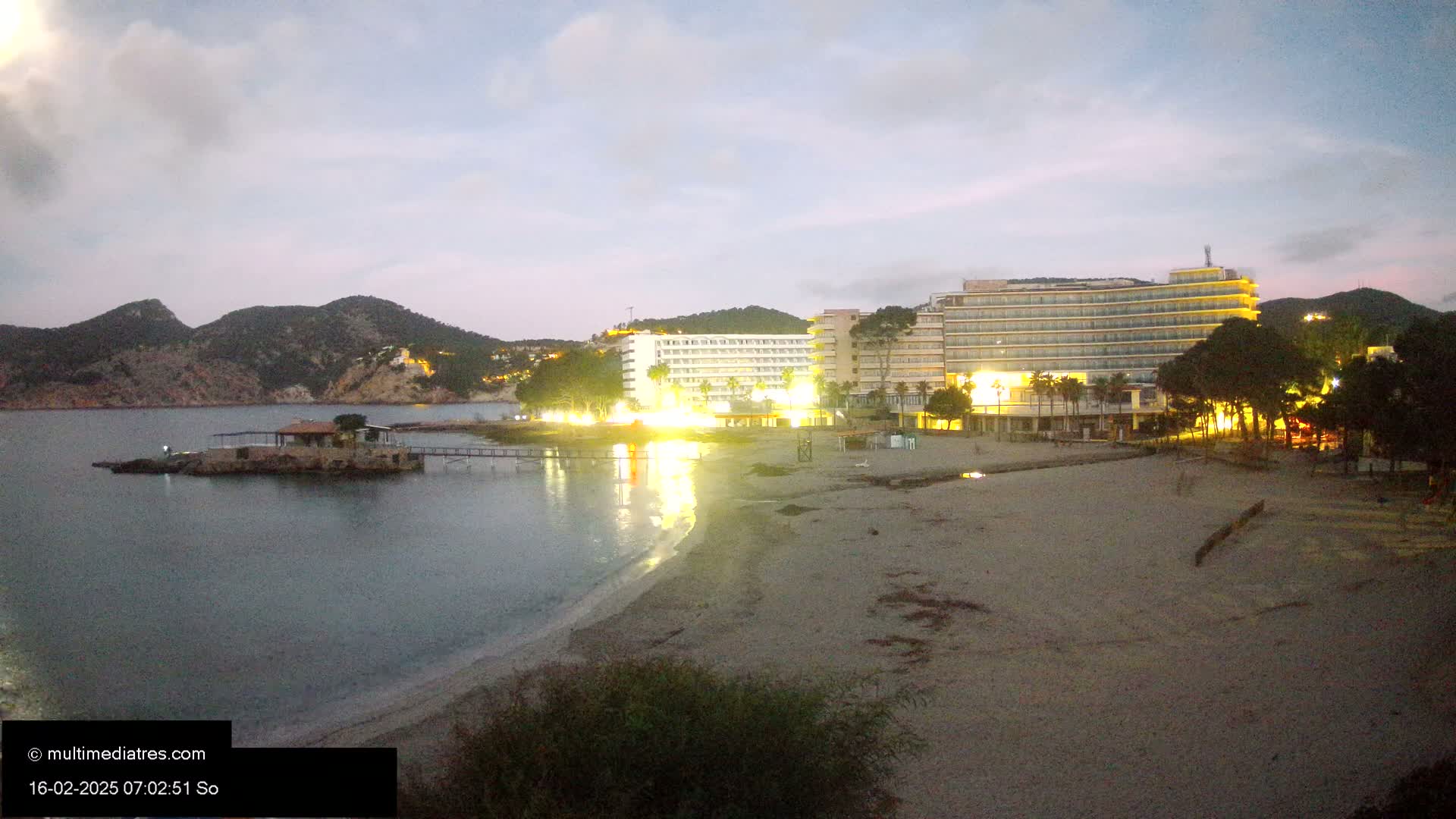 A sandy beach at twilight, with a large hotel illuminated and a small island with a structure in the calm water, backed by dark hills.