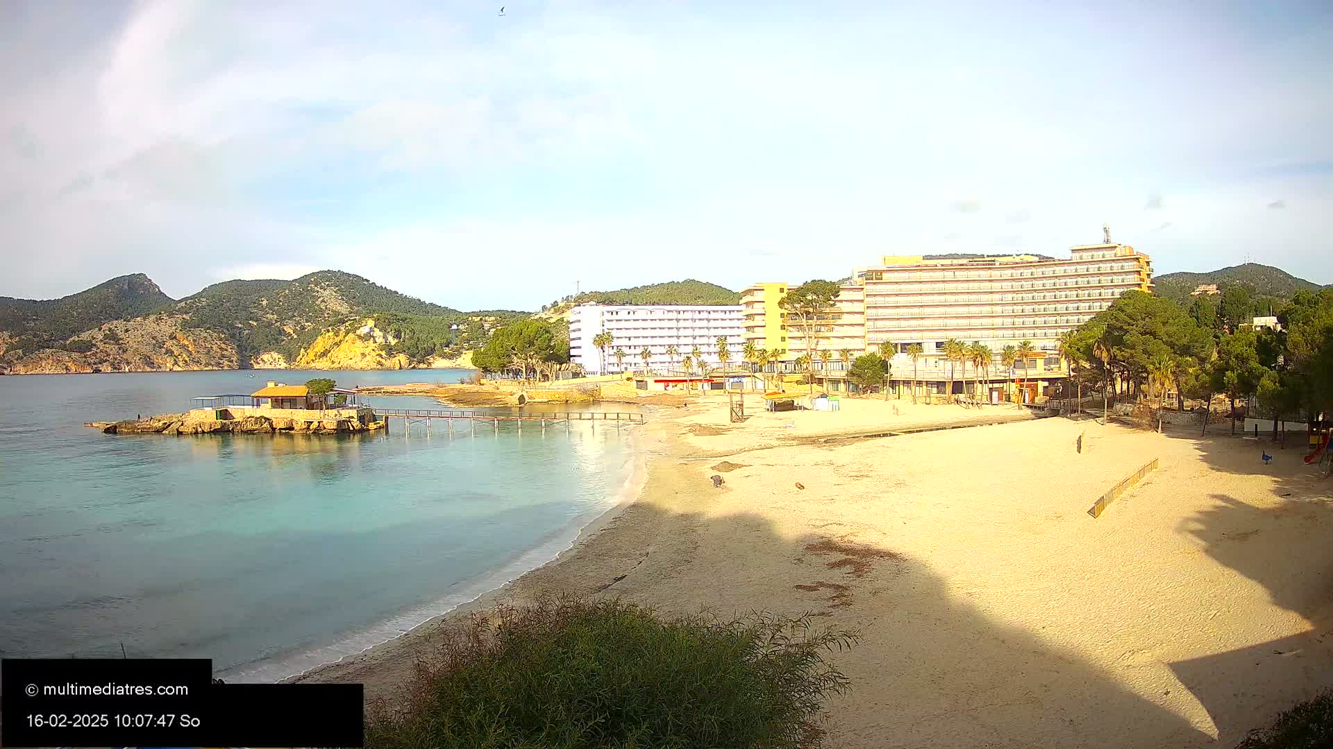 A sandy beach with calm, clear turquoise water laps its shore,  backed by hotels and a small island with a building;  hills rise in the background under a partly cloudy sky.